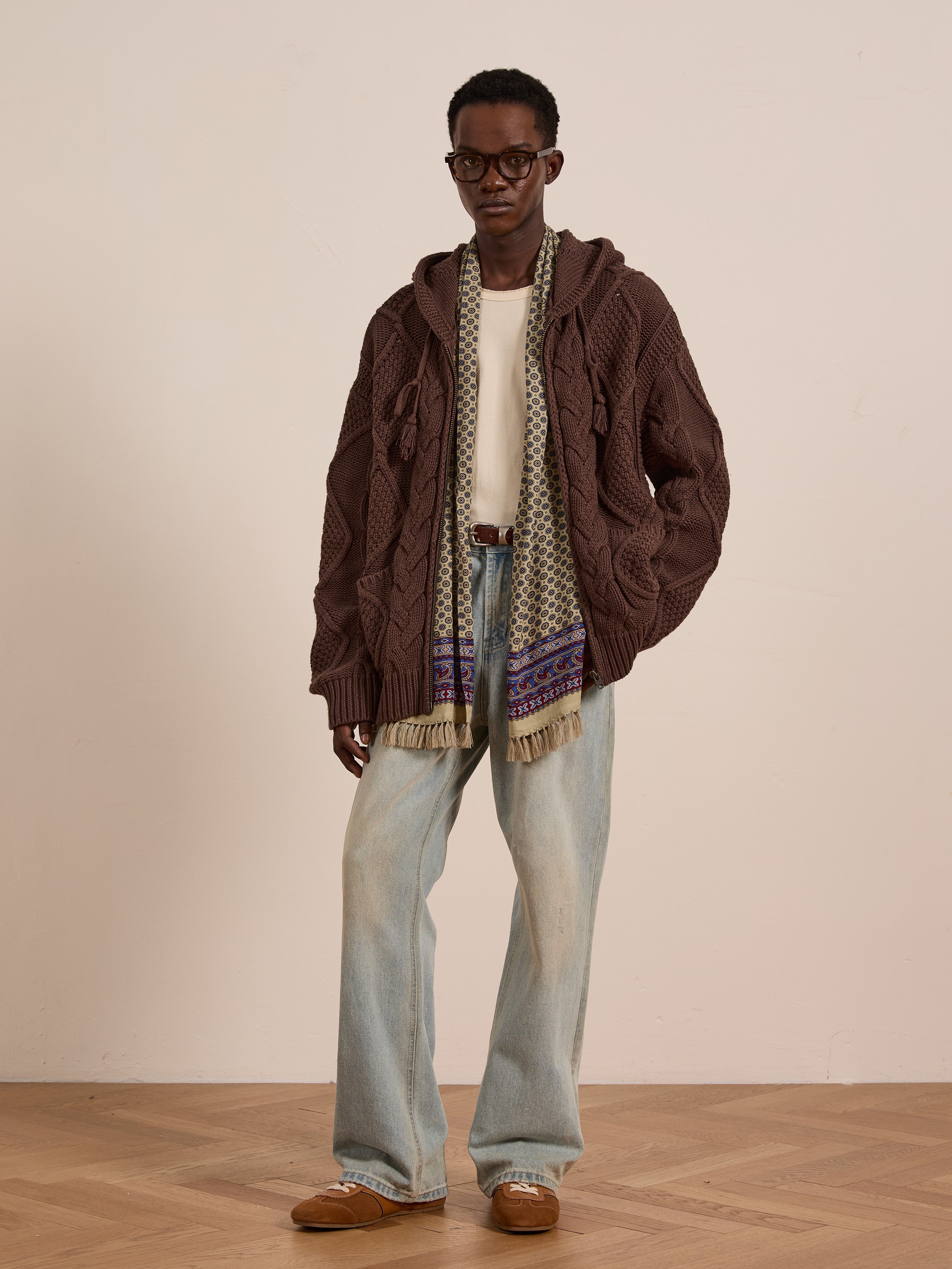 A person wears the FOUND Zip Up Cable Knit Hoodie in brown, paired with a cream shirt, light blue jeans, patterned scarf, and brown shoes while standing on a wooden floor against a plain background.