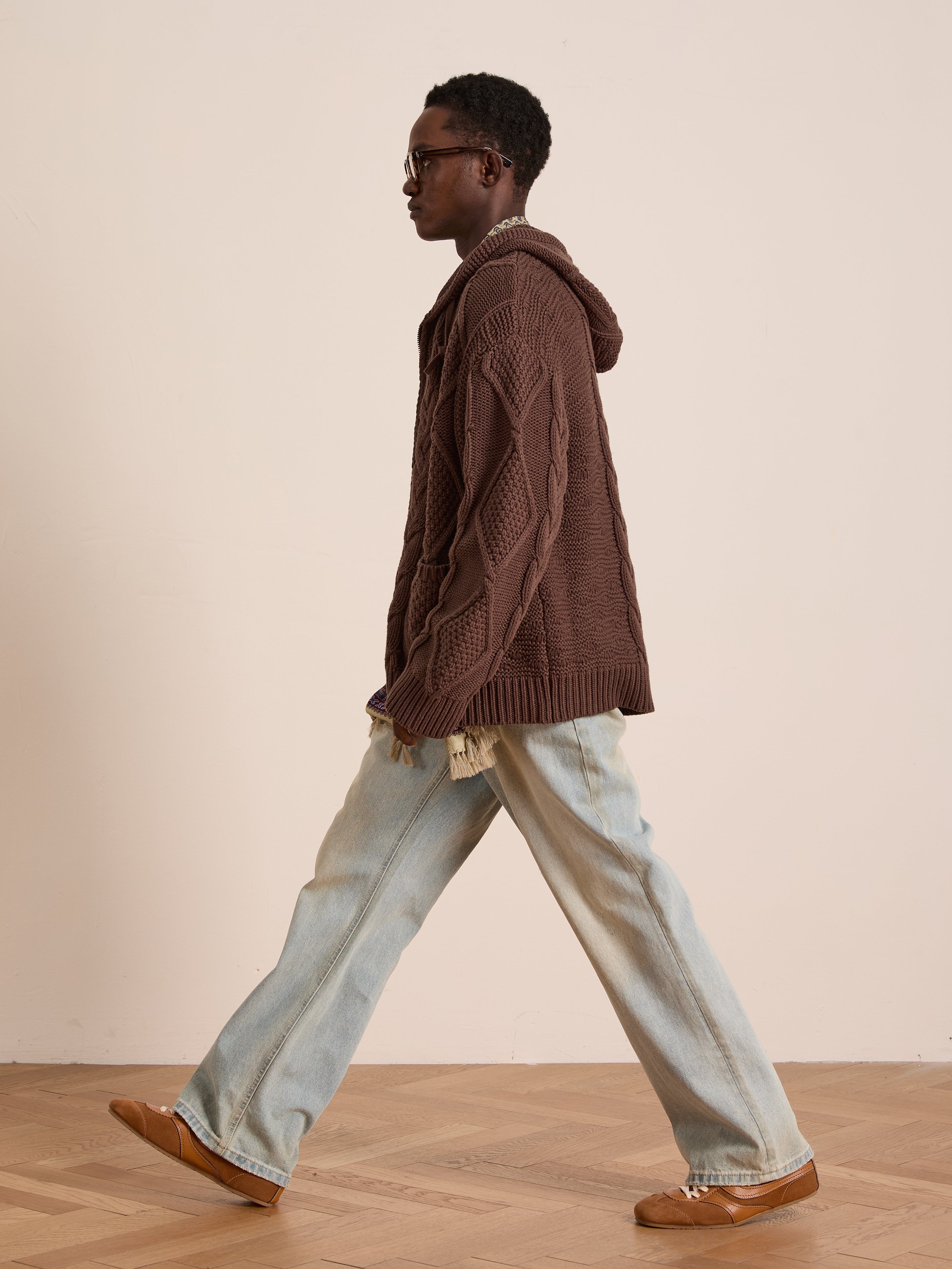 A person wears the FOUND Zip Up Cable Knit Hoodie (brown, unisex relaxed fit) with light blue wide-leg jeans and brown shoes, walking indoors on a wooden floor against a plain beige wall.
