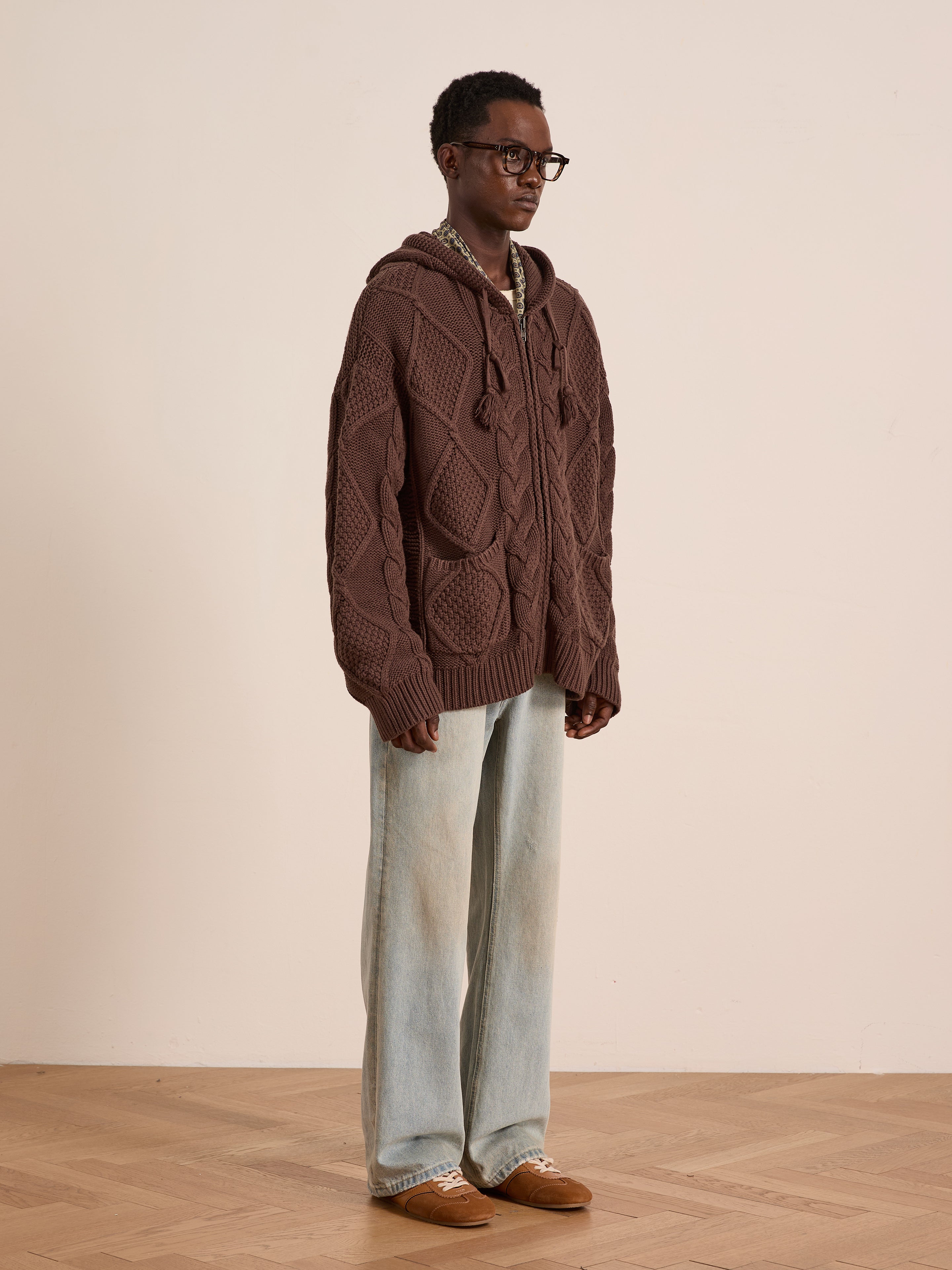 A person wearing black glasses, the FOUND Zip Up Cable Knit Hoodie (unisex, relaxed fit), light blue wide-leg jeans, and brown shoes stands against a plain beige wall on a wooden floor.