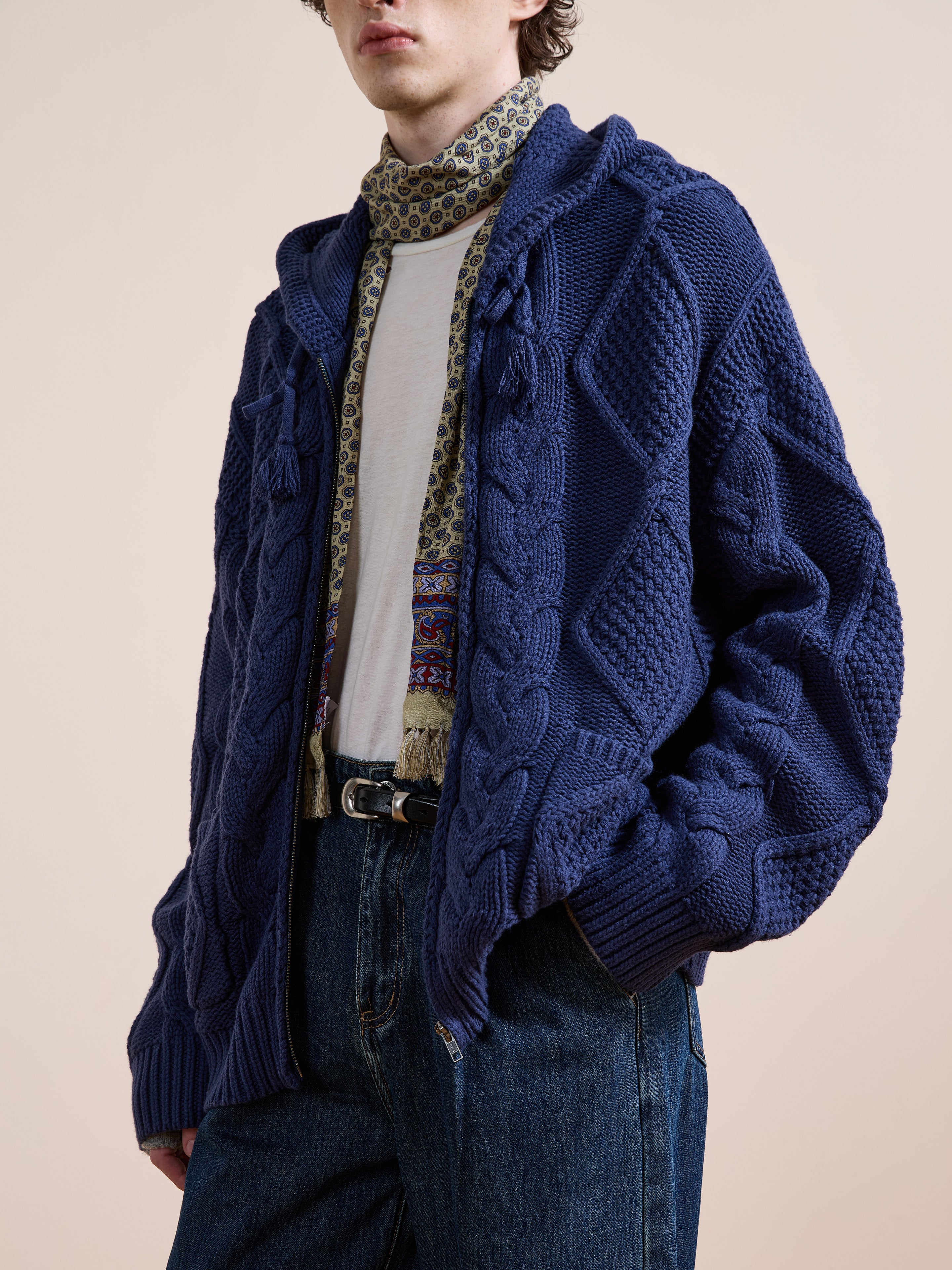 Someone wearing the Found Zip Up Cable Knit Hoodie in textured blue over a white shirt, with a patterned scarf and blue jeans, stands against a plain beige background.
