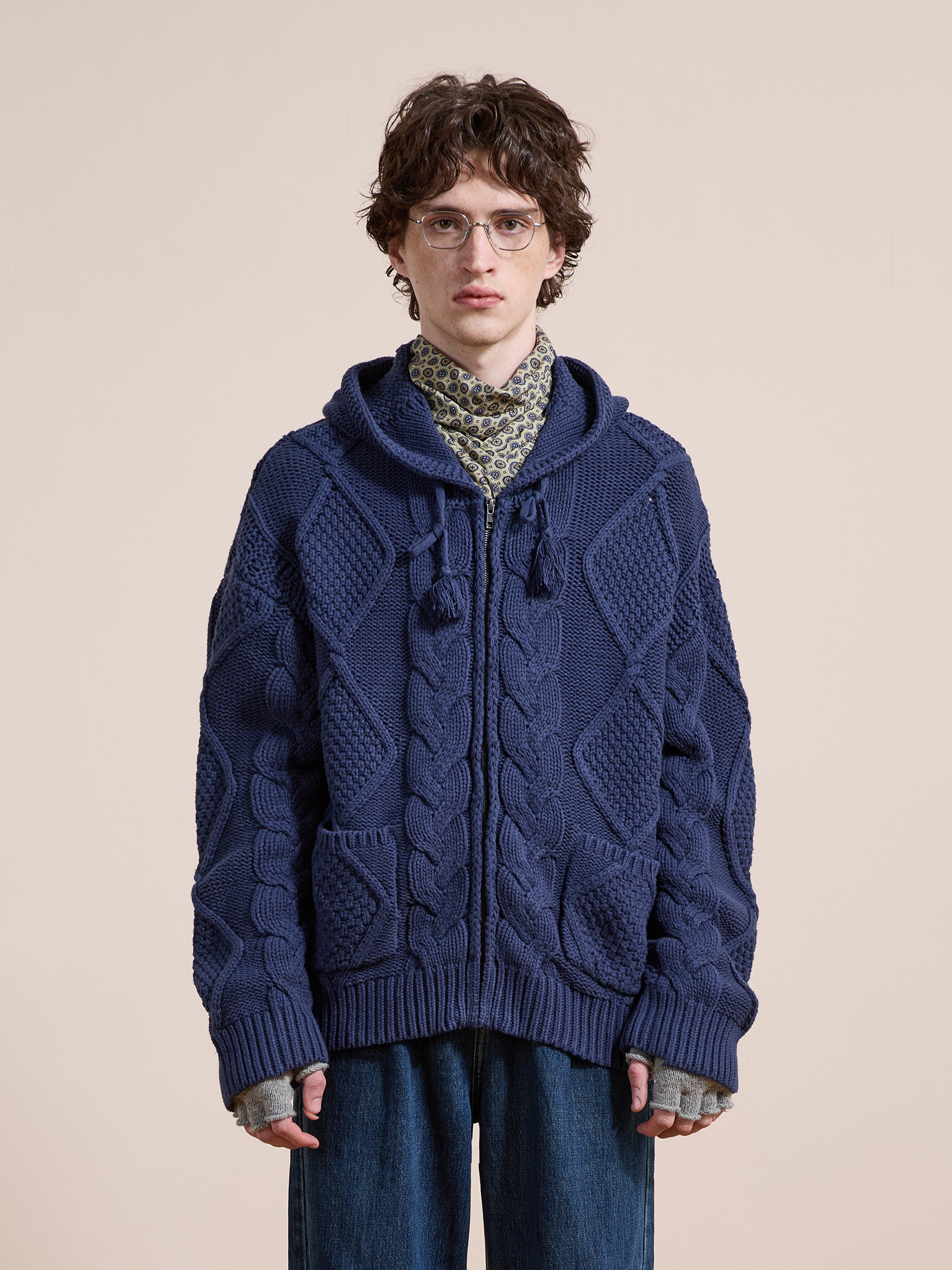 Wearing the Found Zip Up Cable Knit Hoodie in textured navy blue over a patterned shirt and loose denim jeans, a person with glasses stands against a plain beige background.
