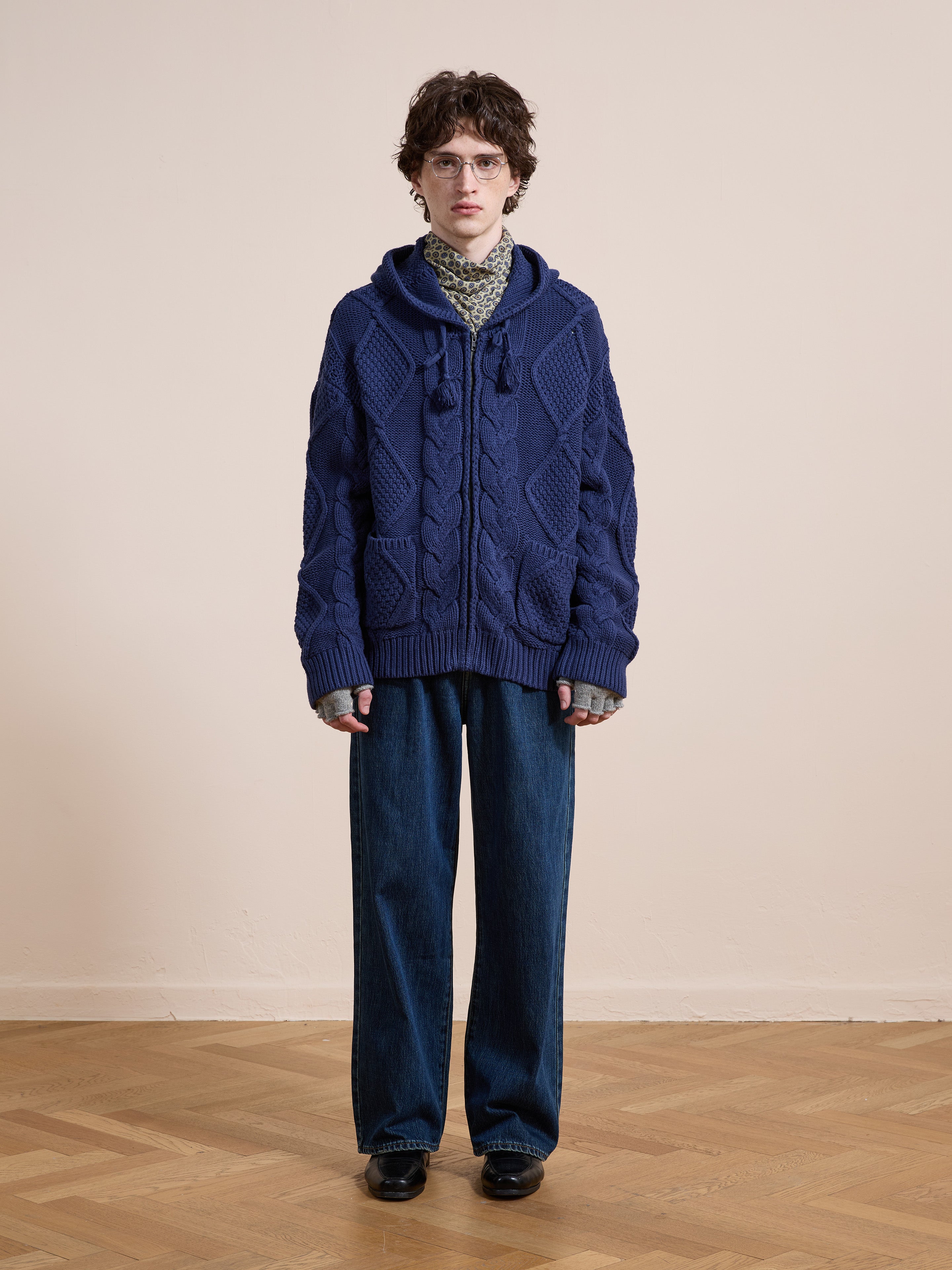 A person stands indoors on a wooden floor, wearing the Found Zip Up Cable Knit Hoodie in blue, wide-leg jeans, black shoes, and glasses against a neutral background.
