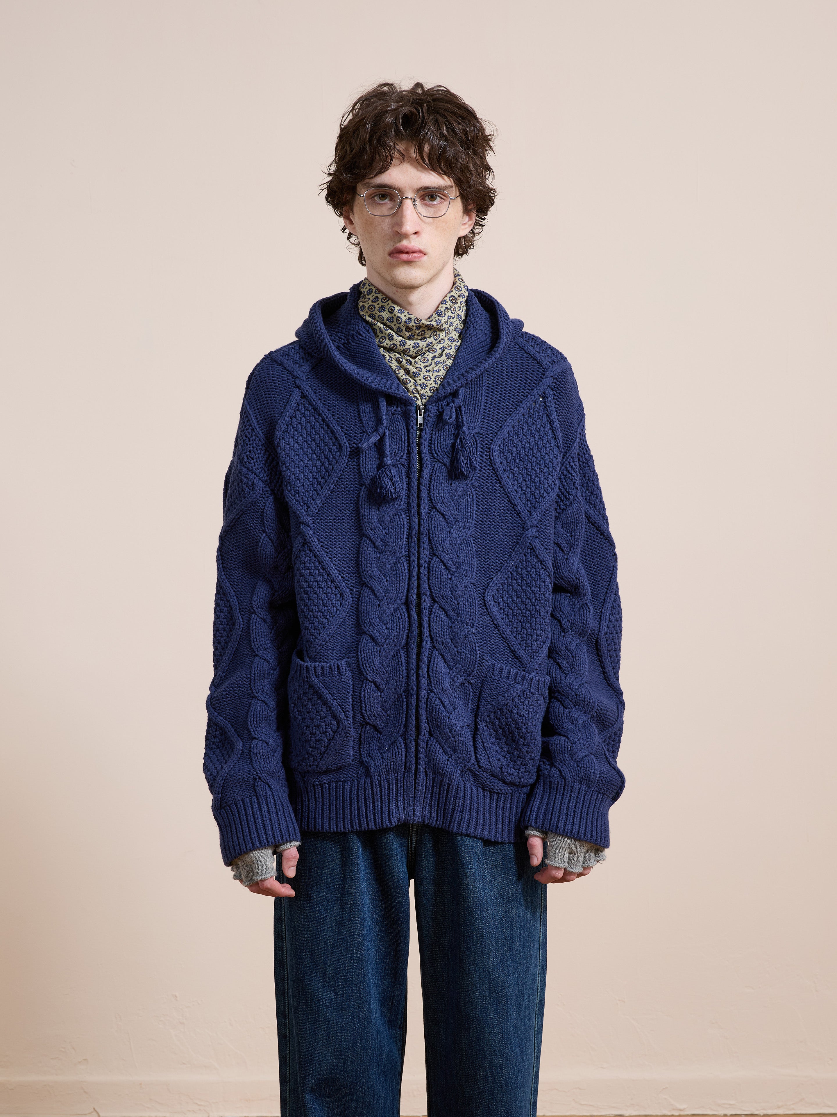A person wearing glasses, a patterned scarf, a chunky blue Found Zip Up Cable Knit Hoodie, and loose blue jeans stands facing the camera against a plain beige background.
