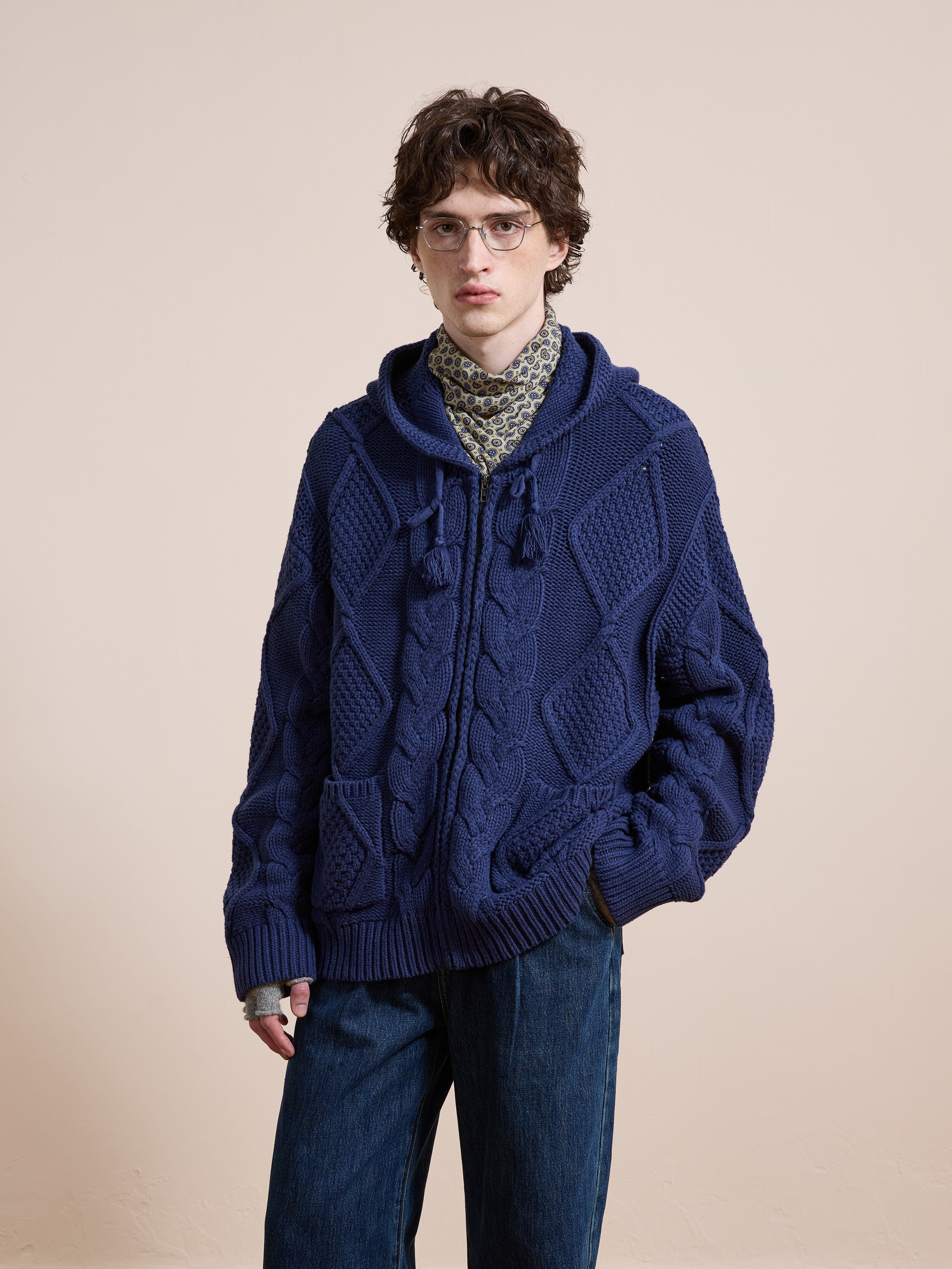 A person wears the Found Zip Up Cable Knit Hoodie—chunky blue with pockets and tassels—over a patterned shirt and glasses, standing against a plain beige background.