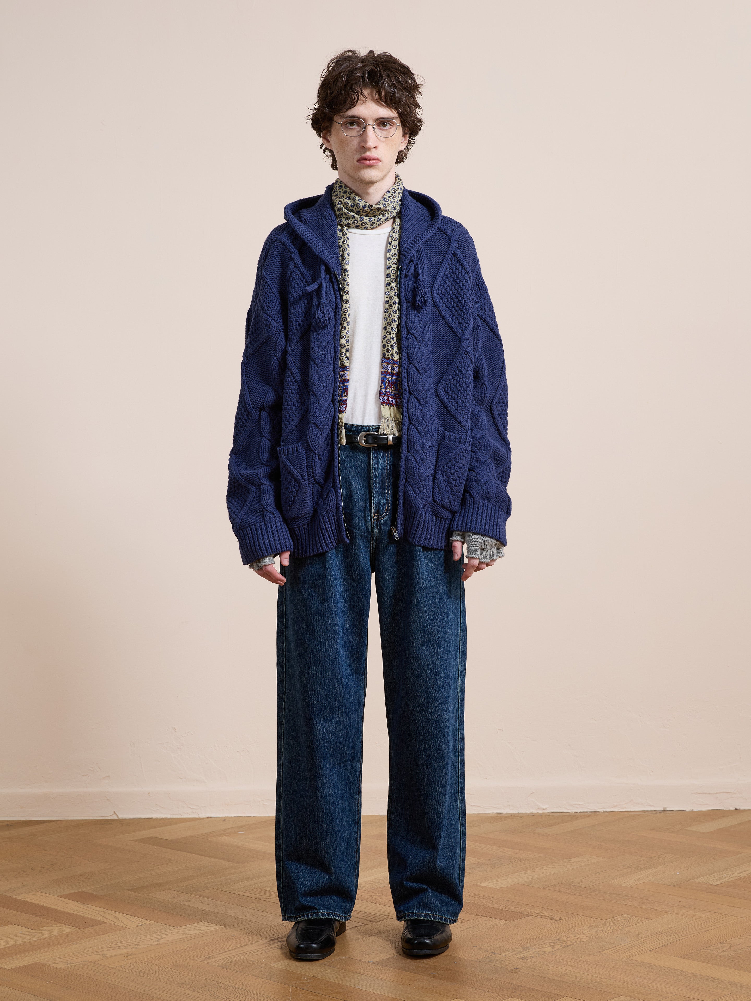 Indoors, a person wears the Found Zip Up Cable Knit Hoodie with a white shirt, patterned scarf, wide-leg jeans, and black shoes on a wooden floor against a plain light wall.