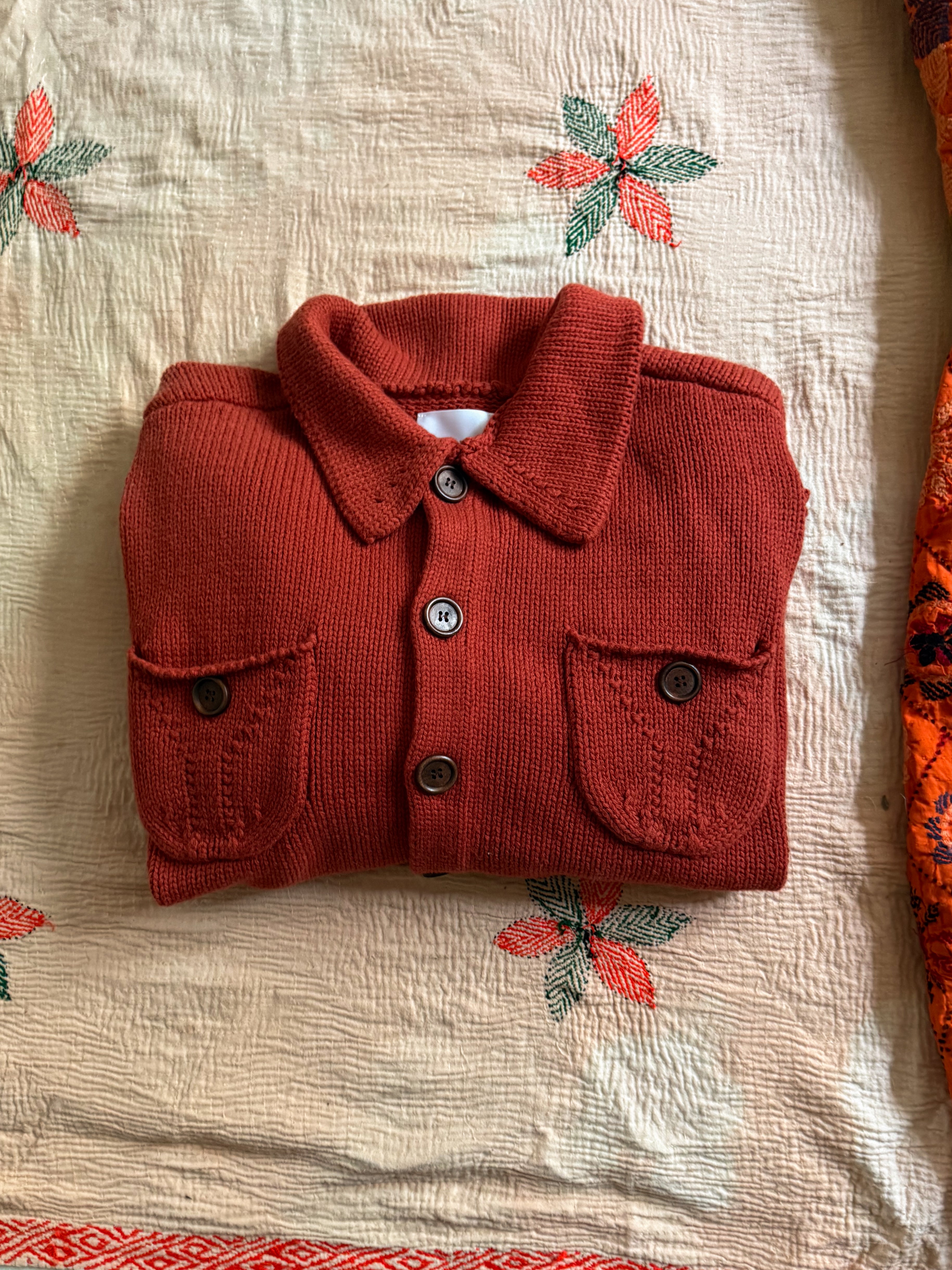 The Hunter Knit Collared Cardigan by FOUND is a folded red pocket cardigan with front buttons, crafted in mid-weight knit featuring two pockets. It's elegantly placed on a floral-patterned fabric backdrop, ideal for transitional layering.