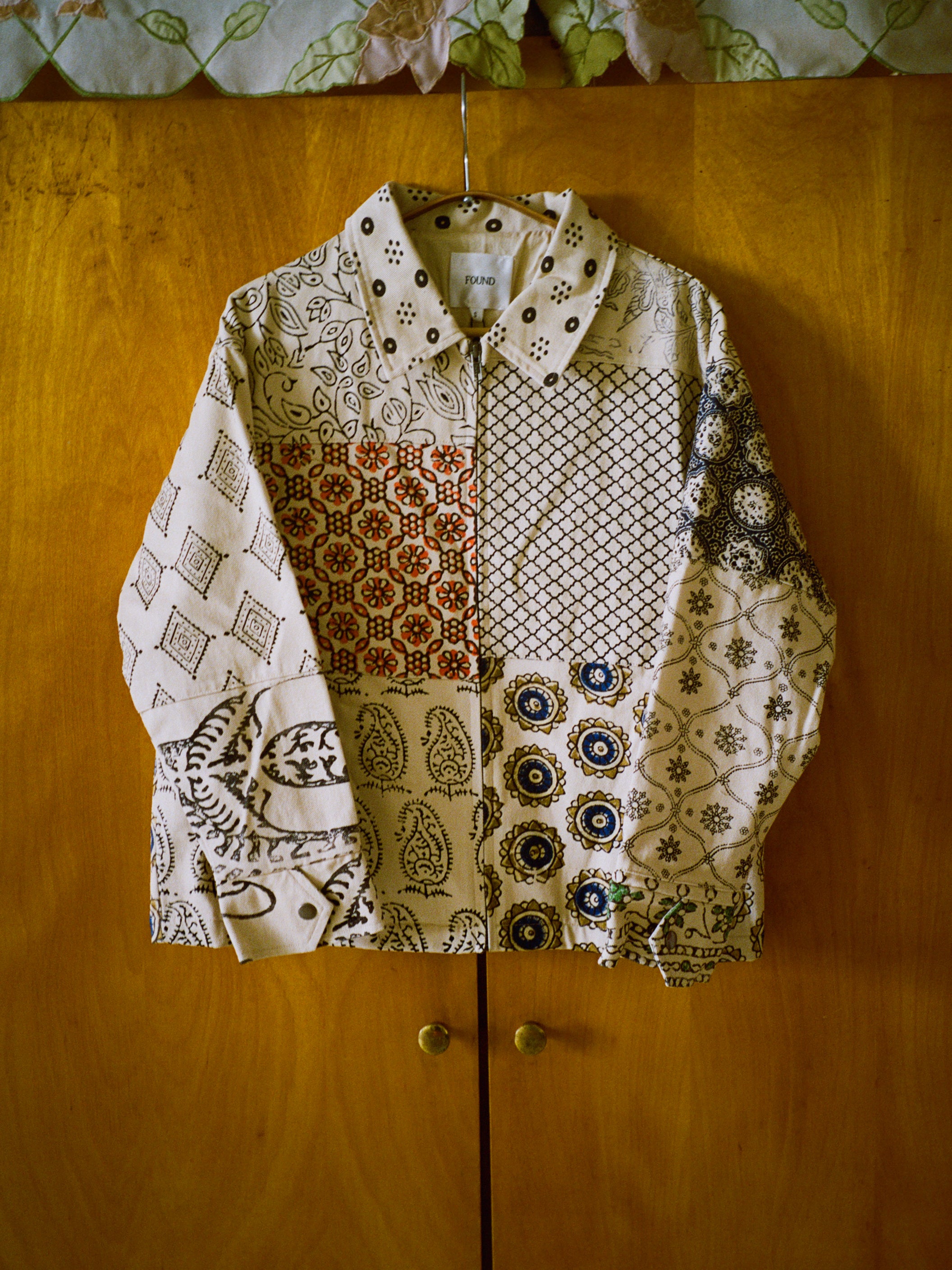 The FOUND Paisley Mosaic Work Jacket, a unisex long-sleeve cotton twill shirt with vibrant patterns and colors, hangs on a wooden door.