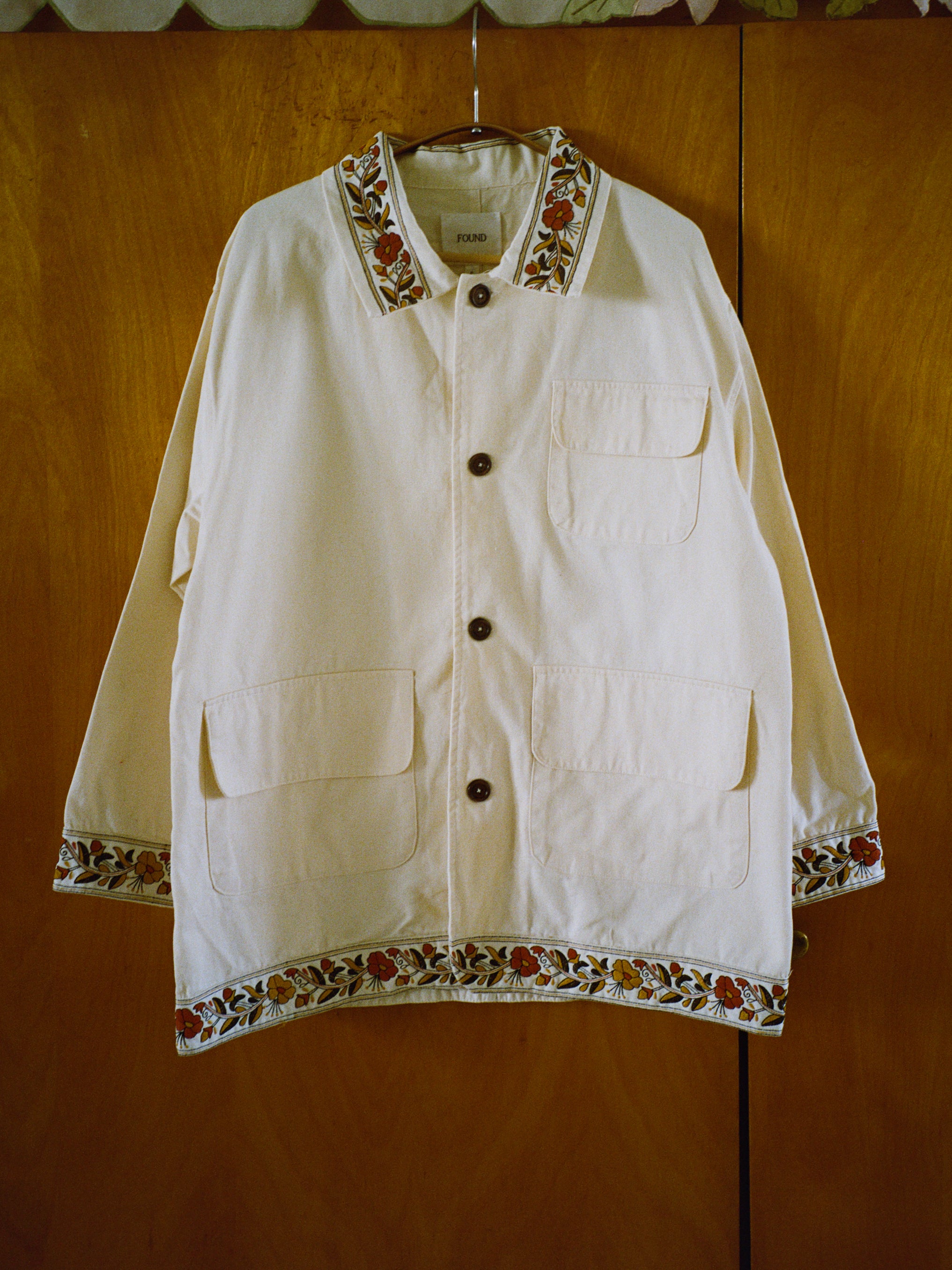 A cream-colored Kantha Floral Work Jacket by FOUND, adorned with floral kantha embroidery on the collar, cuffs, and hem, hangs against a wooden background. It features two large pockets and a smaller chest pocket, highlighting cherished heritage quilting techniques.
