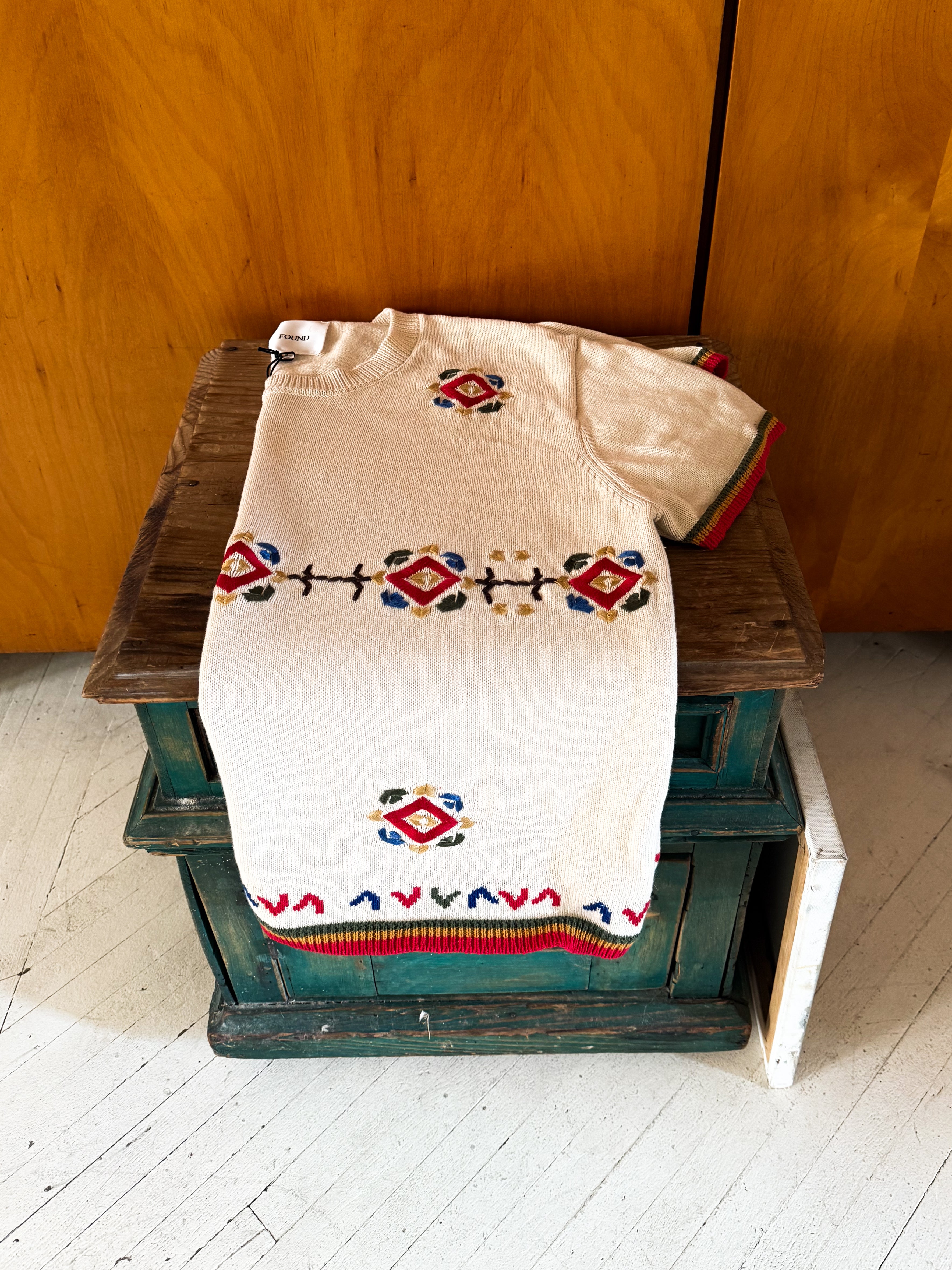 A folded white Knit Flower Petal Tee by FOUND, boasting vintage-inspired charm with jacquard-like floral patterns in red, blue, and green, rests gracefully on a wooden chest.