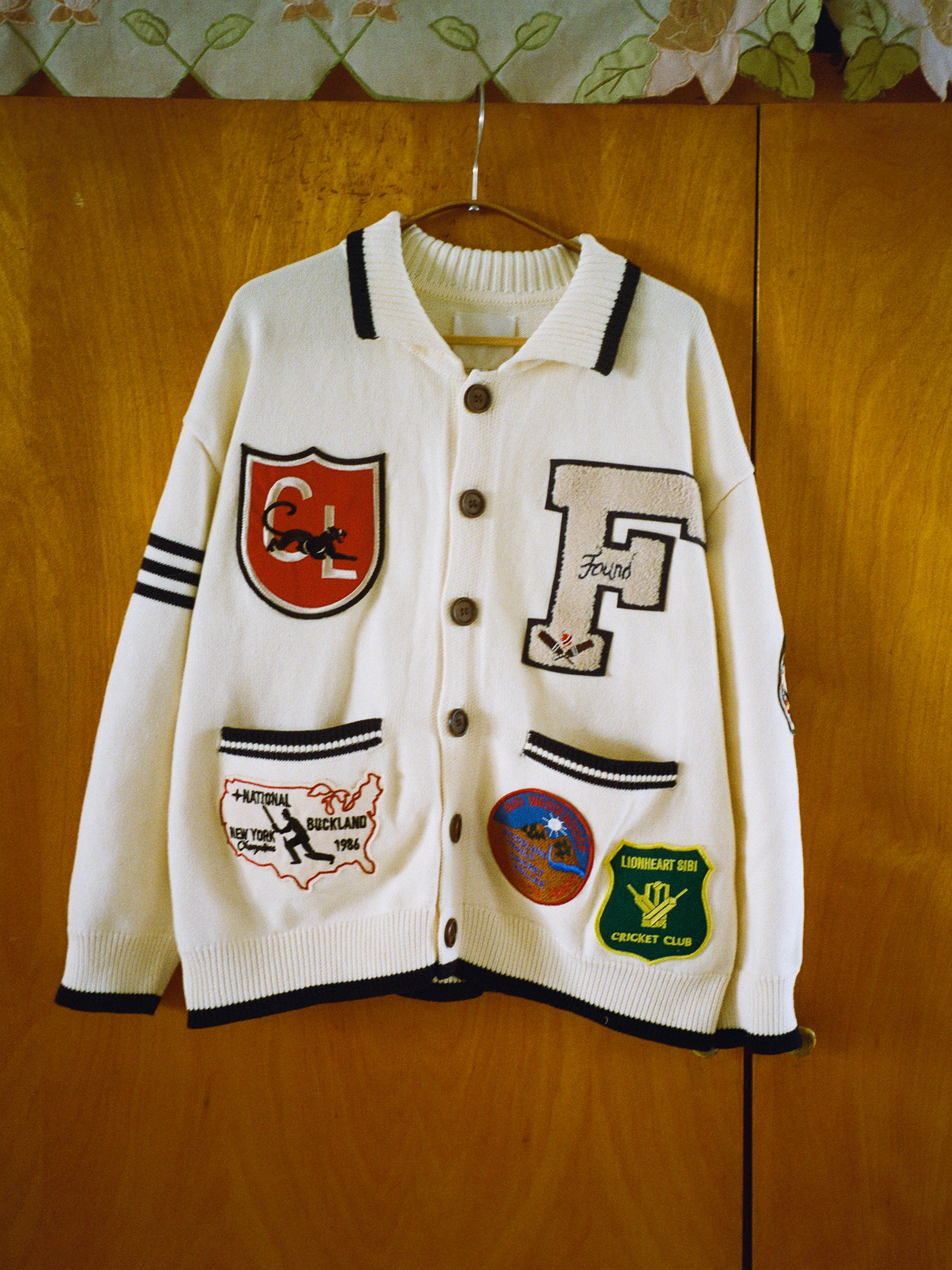 FOUND's York Varsity Patch Collared Cardigan, draped on a wooden surface, evokes collegiate nostalgia with its collection of letters, logos, and maps.