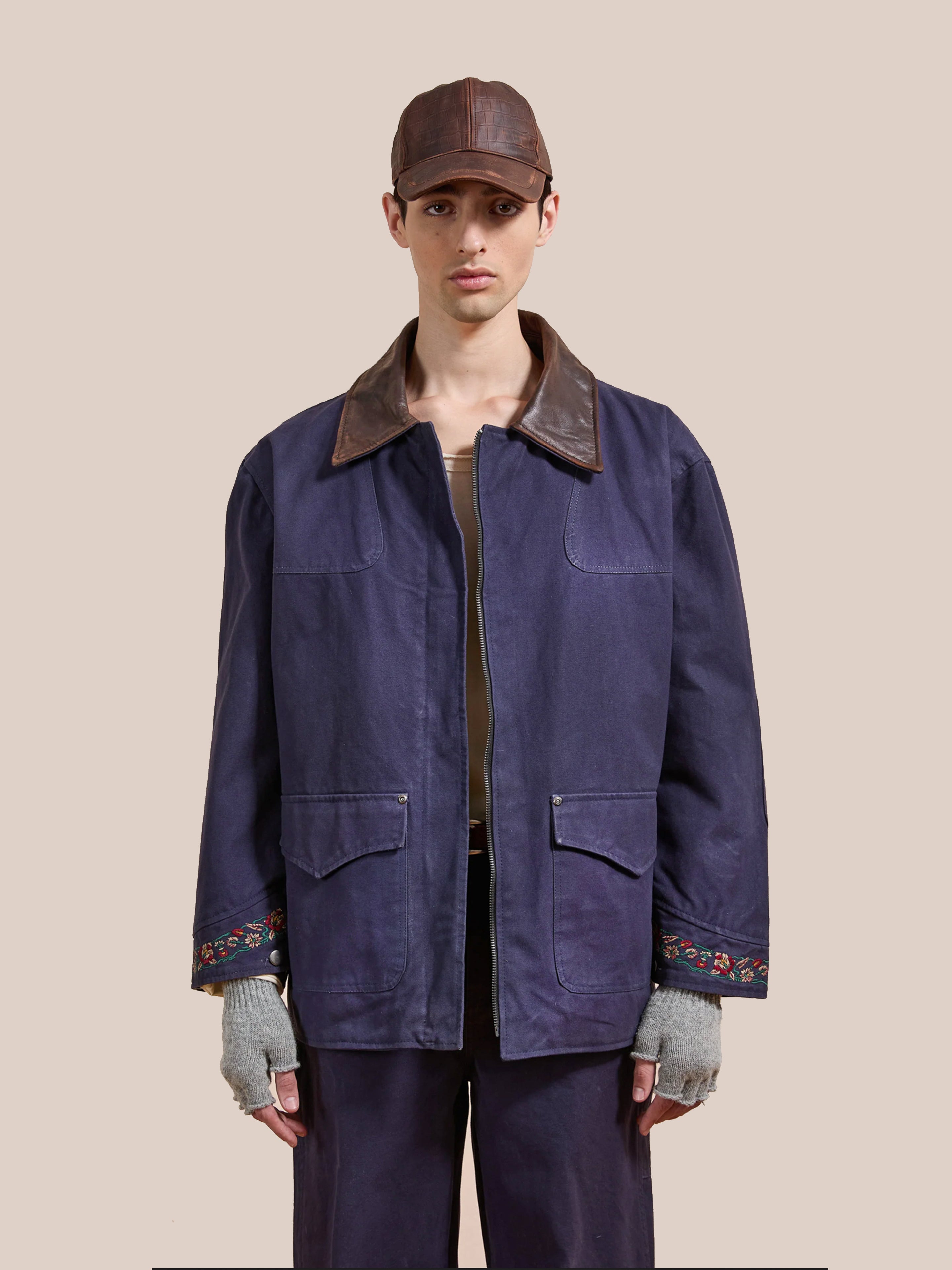 A person wears Found's Canvas Distressed Barn Jacket—navy with a distressed leather collar, oversized fit, and embroidered cuffs—paired with gray fingerless gloves and matching pants, standing against a plain beige background.