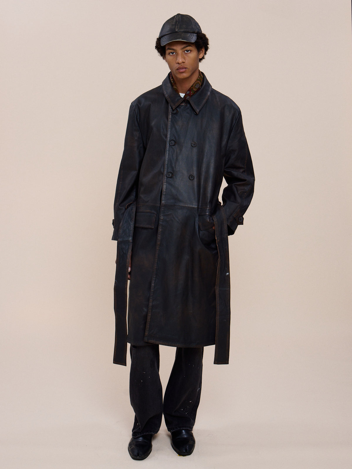 A person in the FOUND Belted Leather Trench Coat with a matching cap and black pants stands against a plain beige background.
