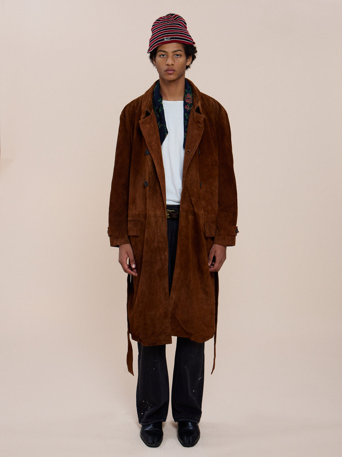 A person stands against a plain background, wearing the FOUND Belted Suede Trench Coat, a white shirt, dark pants, black shoes, and a red striped bucket hat.