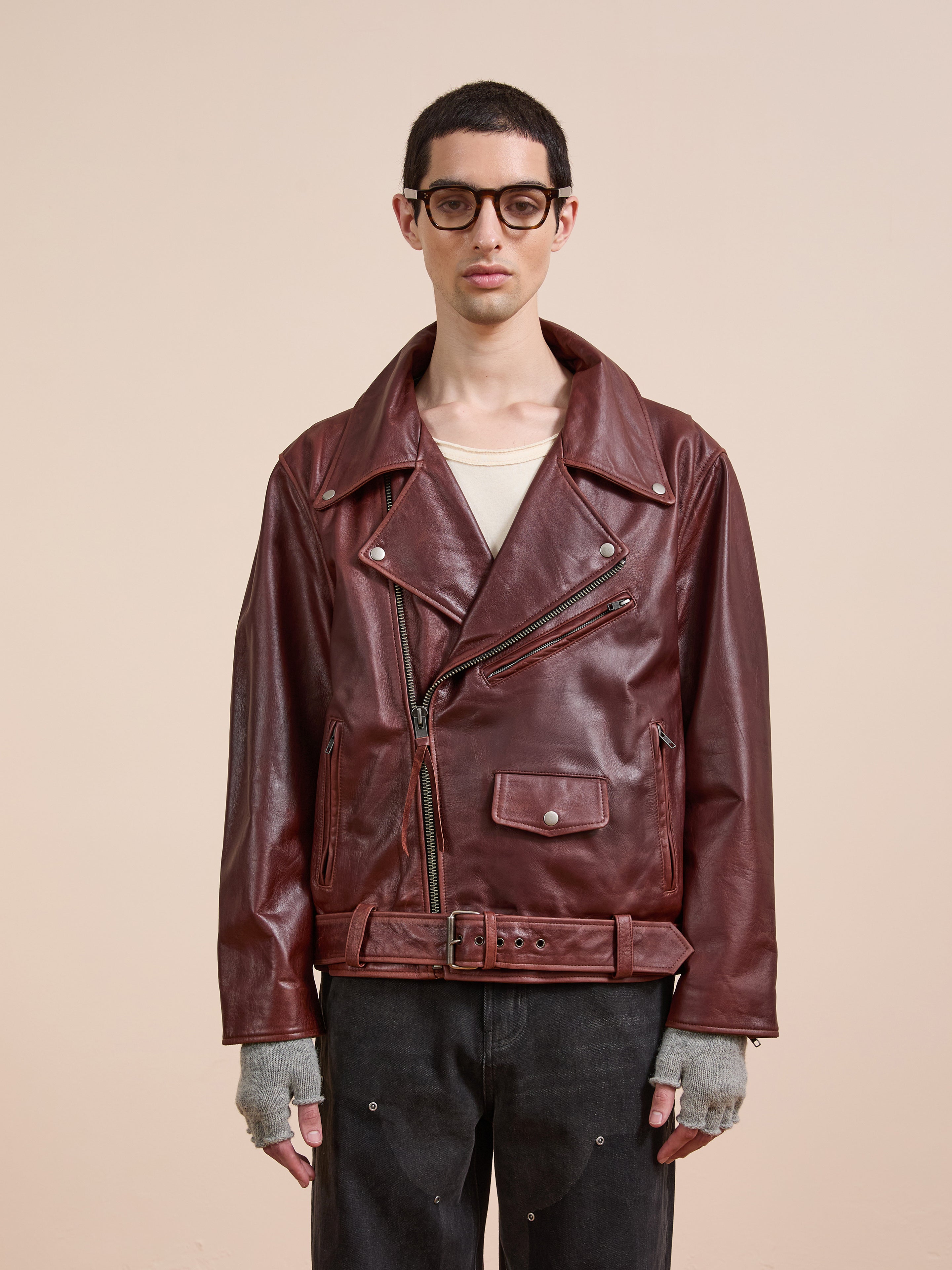 A person wears the FOUND Leather Biker Jacket over a beige shirt, paired with glasses, fingerless gray gloves, and black jeans, standing against a plain light background.