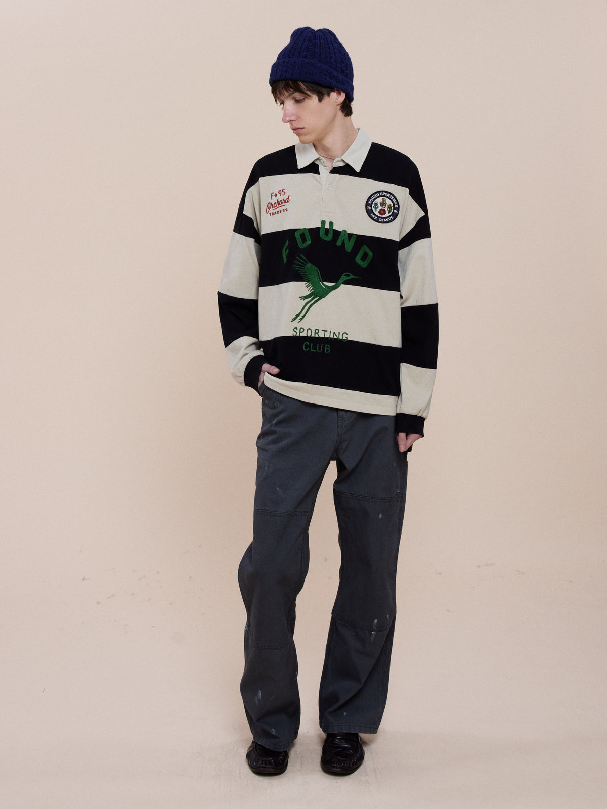 Person in a FOUND Bird Logo Rugby, loose gray pants, blue knit beanie, and black shoes stands against a plain background looking down with hands in pockets.
