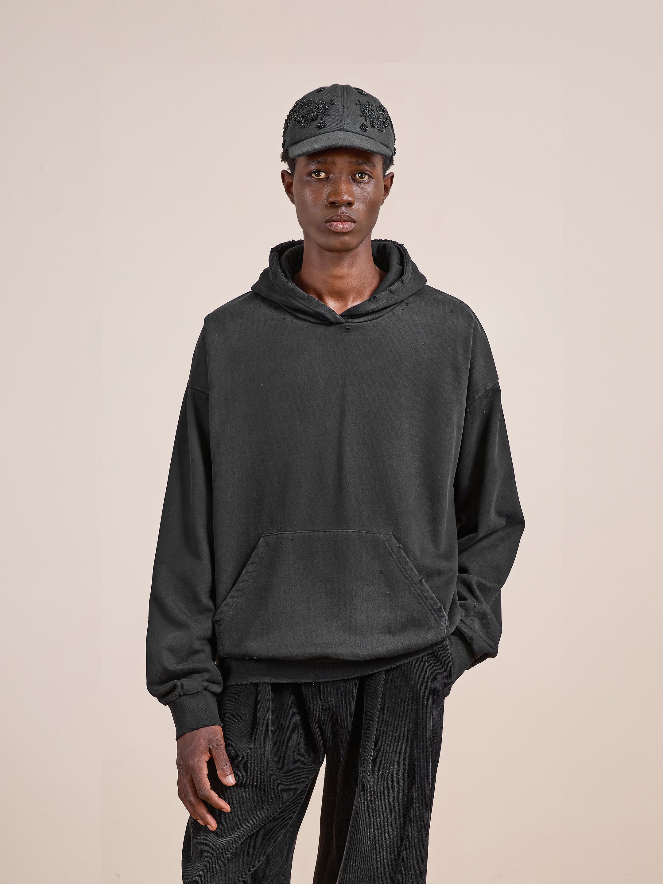 A person wearing the FOUND Distressed Pullover Hoodie in washed black, paired with black pants and a black cap, stands against a plain beige background.