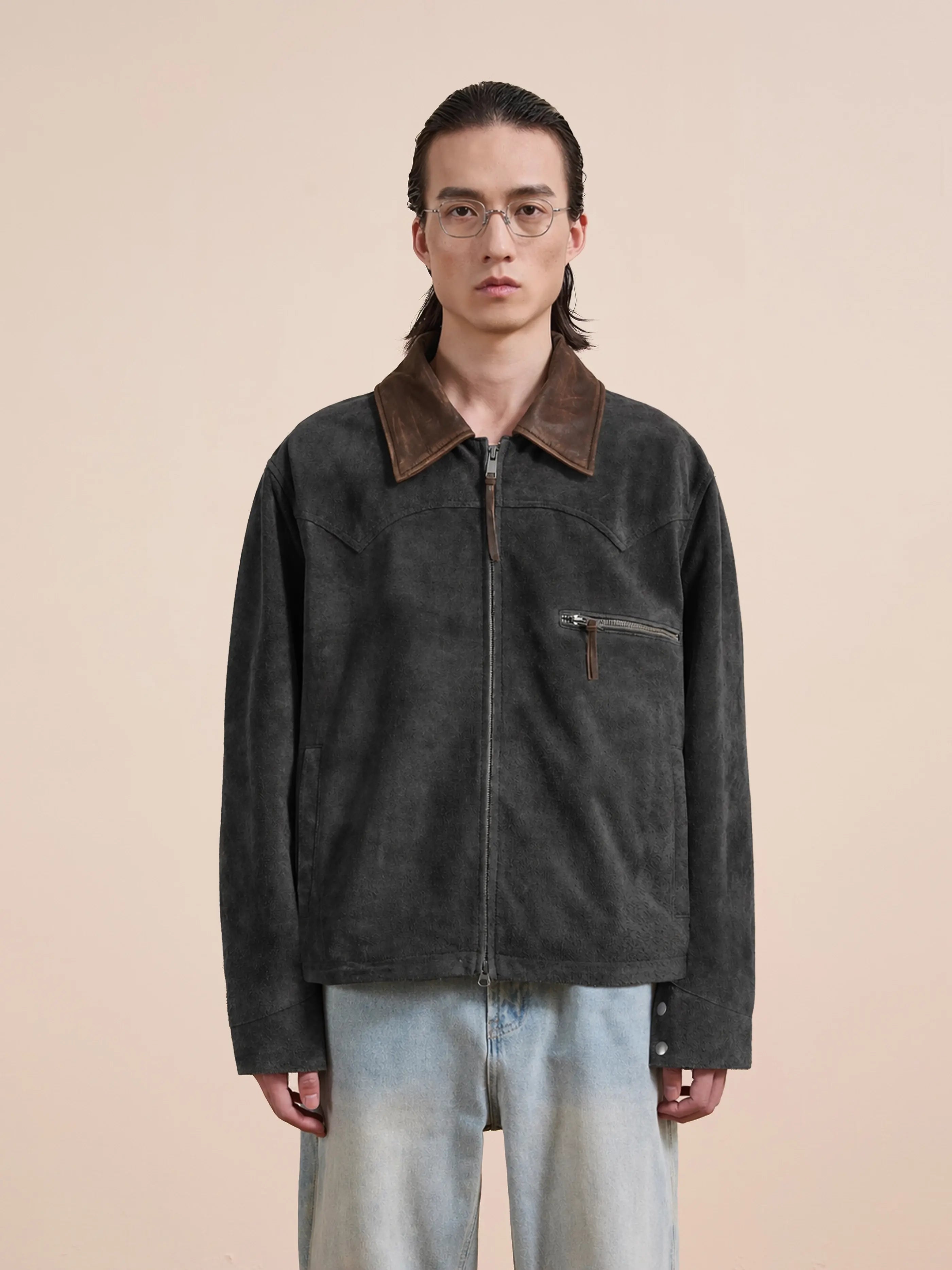 A person with long hair and glasses wears the FOUND Suede Western Work Jacket—vintage black suede with a brown collar, chest pocket, unisex relaxed fit—paired with light blue jeans, standing against a plain beige background.