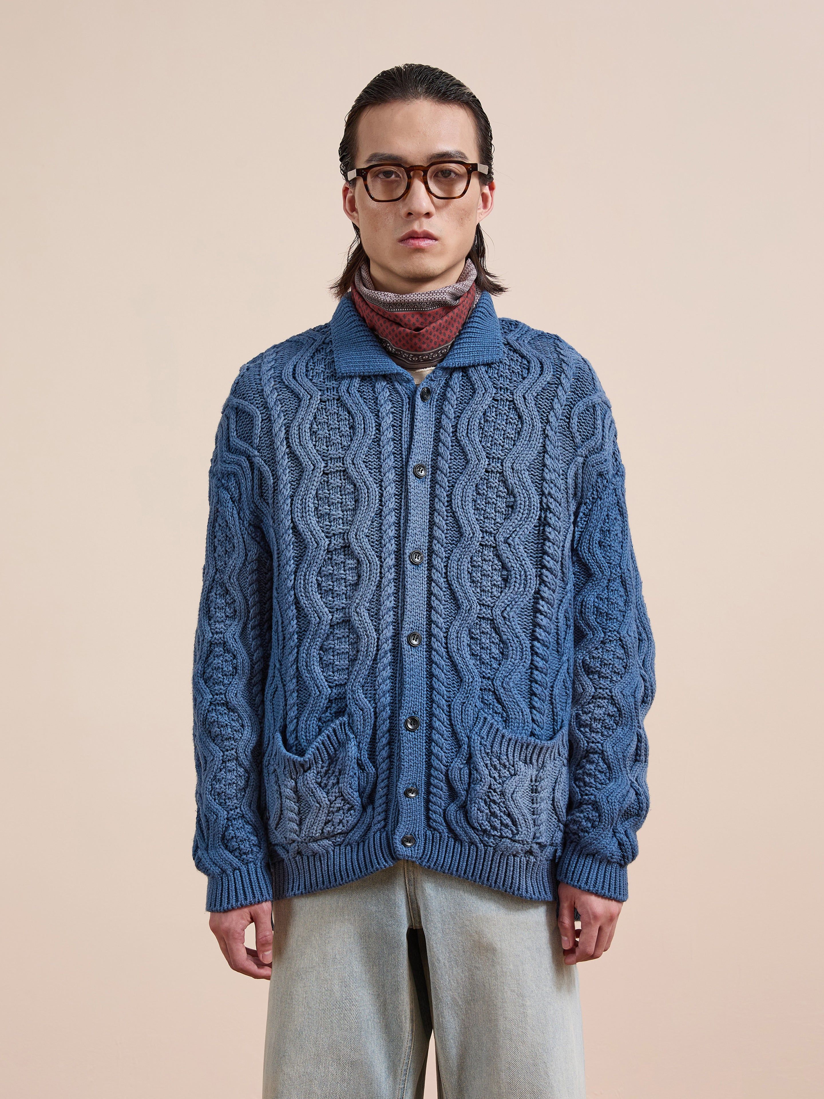 Someone wears Found's Cable Knit Buttoned Cardigan with front pockets, layered over a turtleneck and paired with light blue jeans and glasses, standing against a beige background.