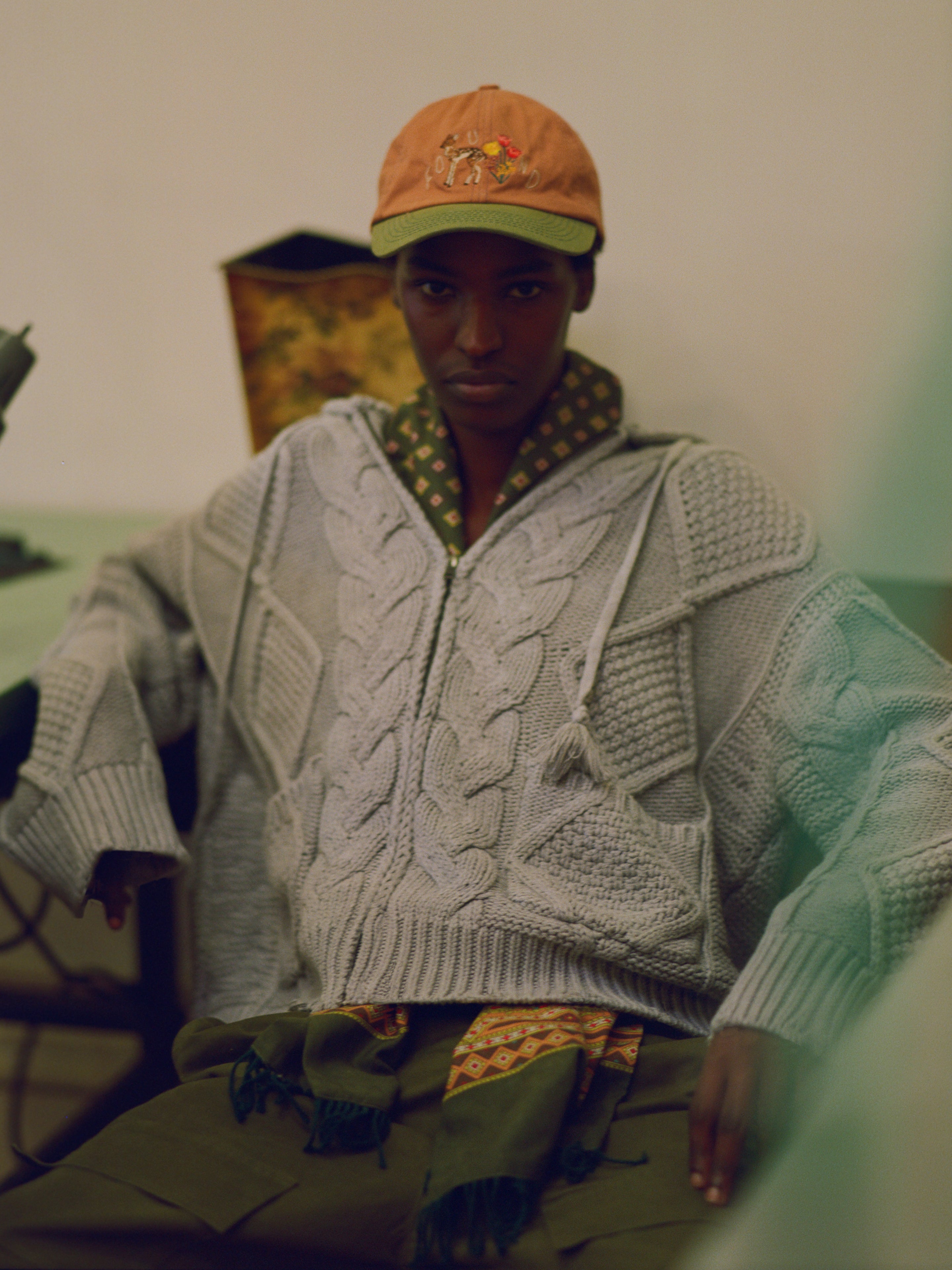 A person wearing a gray Found Zip Up Cable Knit Hoodie with a cozy look and feel, a green patterned shirt, and an orange cap sits with a relaxed posture, hands resting on their knees.