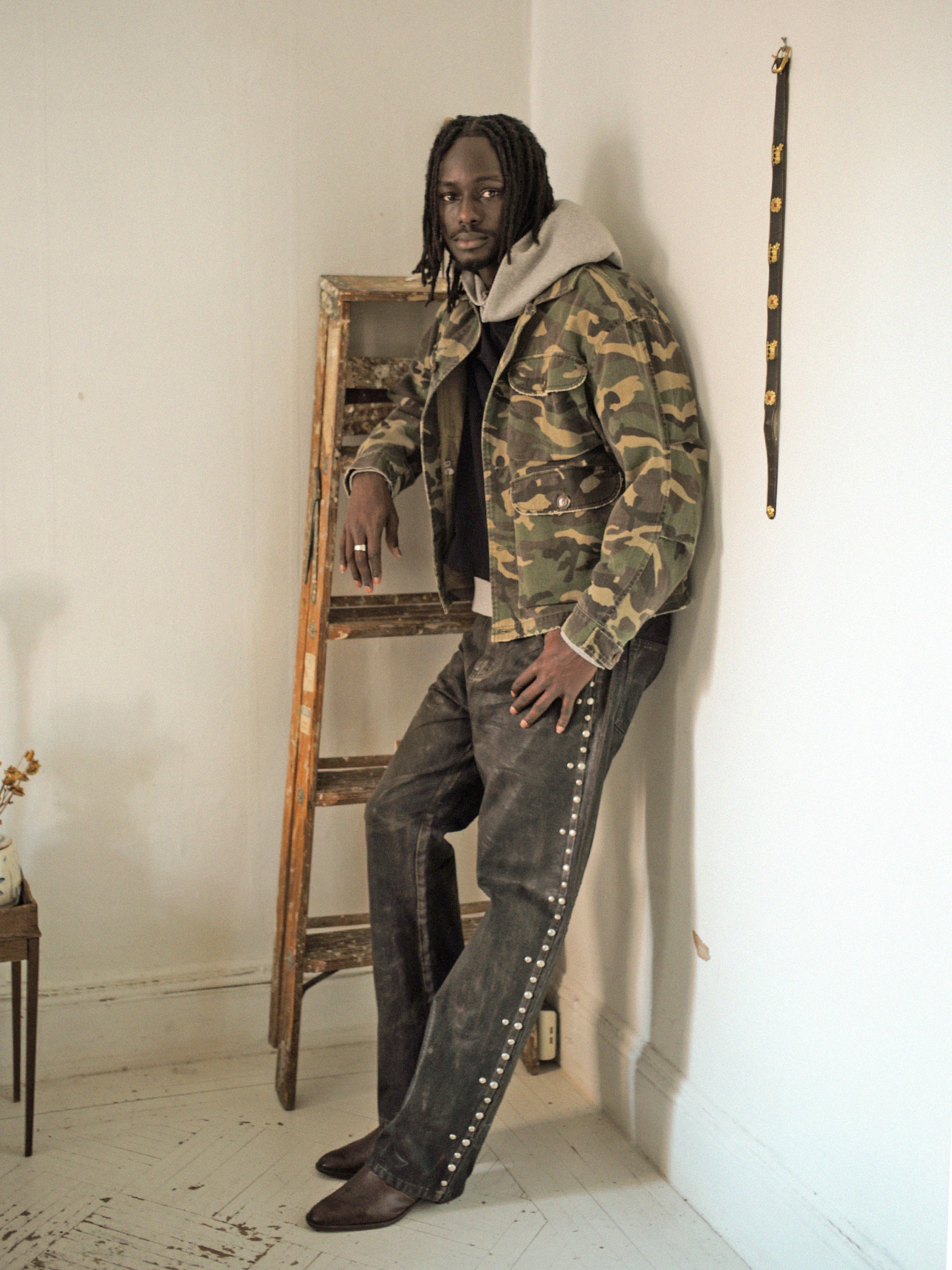 Someone wearing a Camouflage Twill Utility Cargo Jacket by FOUND and dark pants leans against an indoor wooden ladder. A strap with gold details is hanging on the wall beside them.