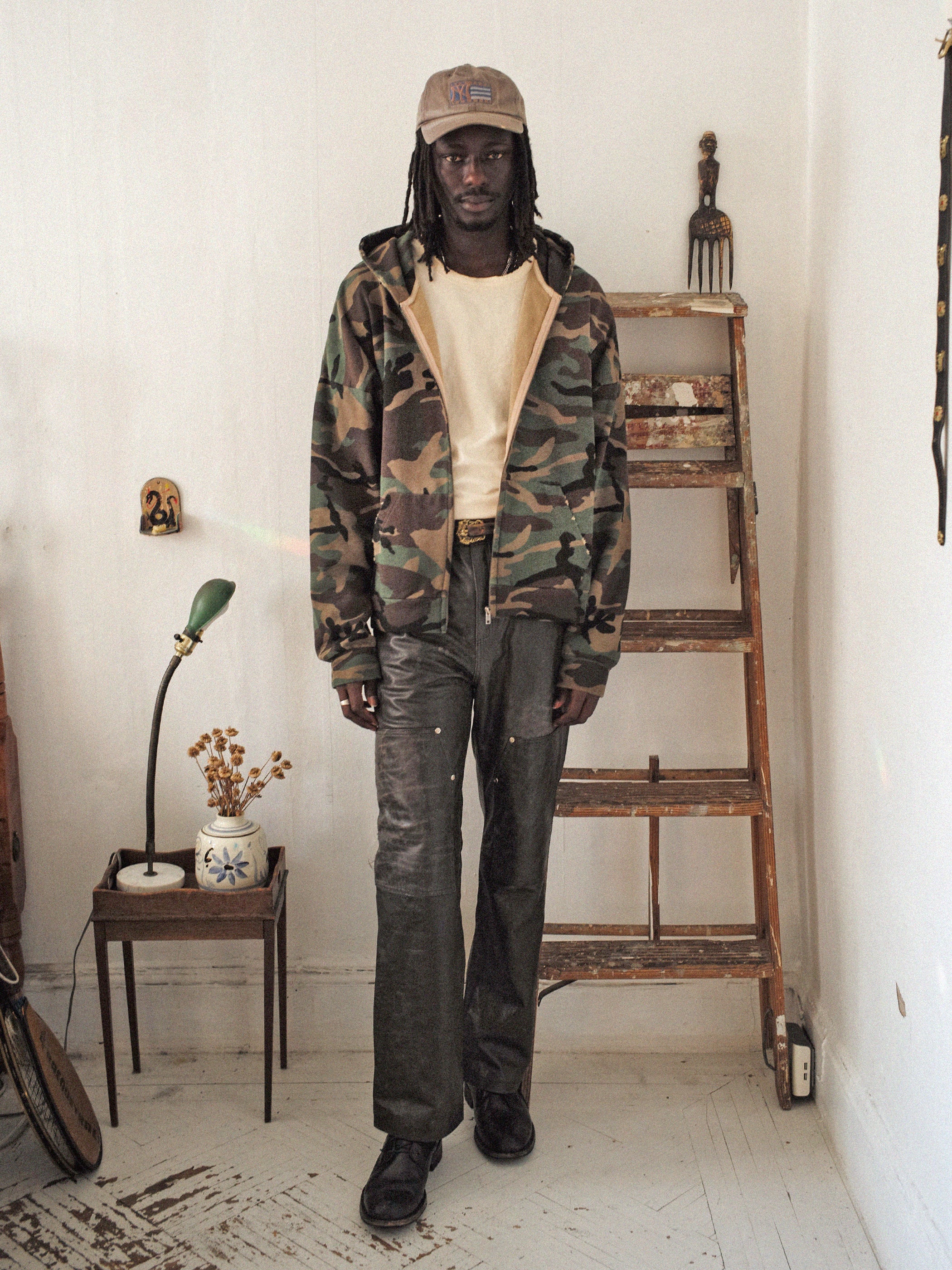 A person wearing a FOUND Washed Camouflage Zip Hoodie, a beige cap, and dark pants stands in a room with a wooden ladder and a small table featuring a vase with flowers, creating a vintage effect.