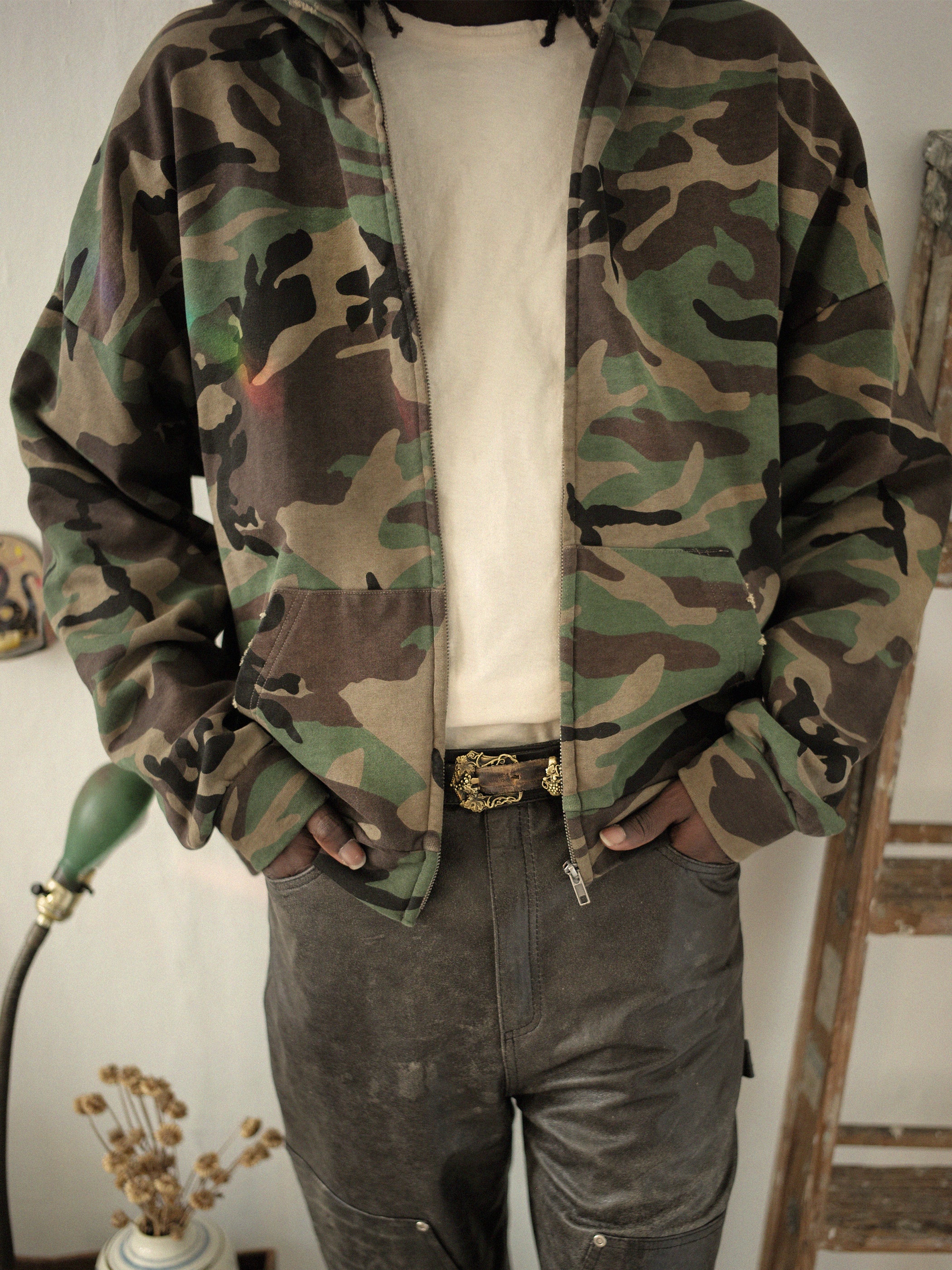 A person wearing a FOUND Washed Camouflage Zip Hoodie and dark pants stands indoors, hands in pockets, giving off a vintage vibe. The backdrop includes a lamp and dried flowers.