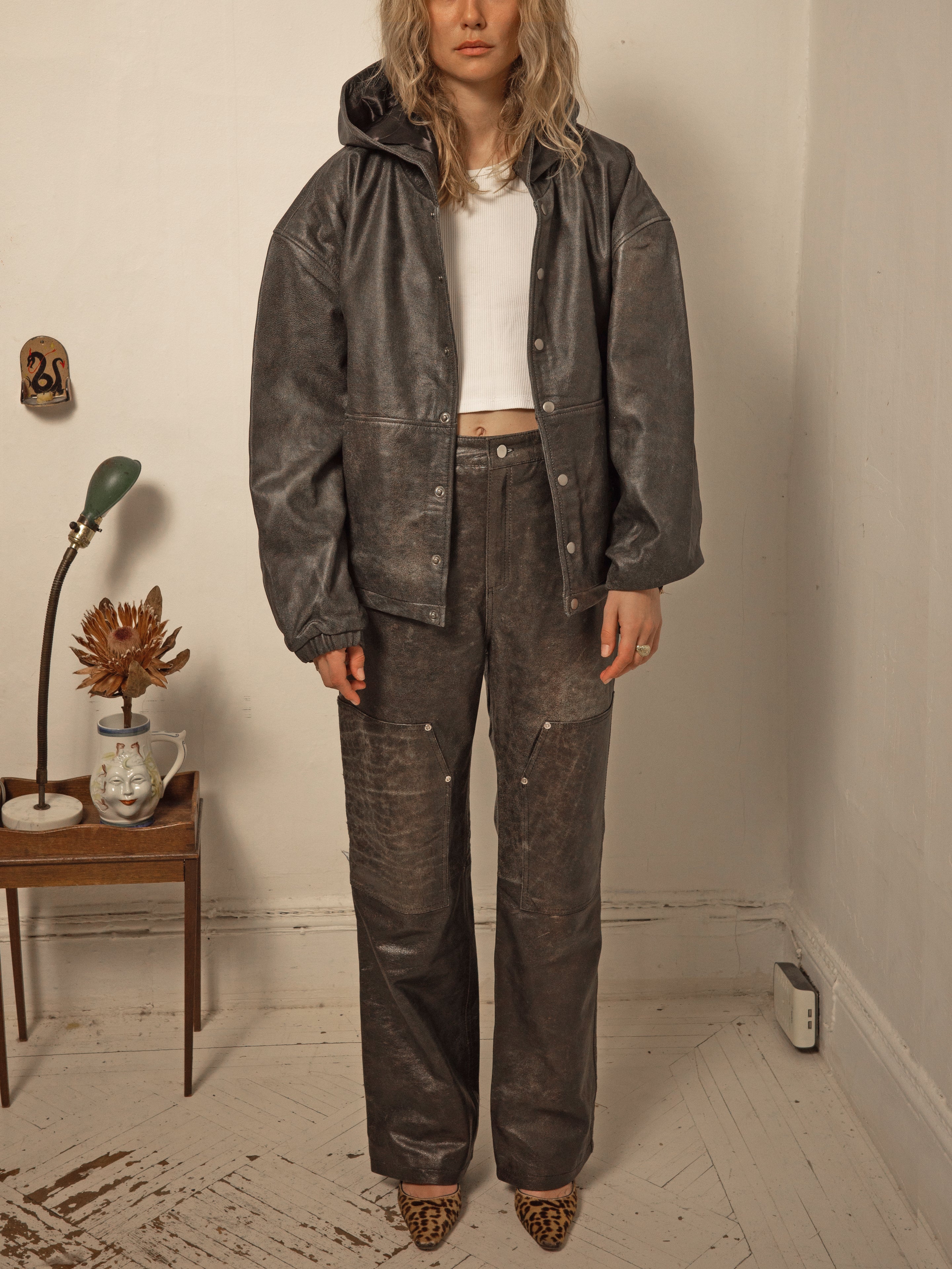 A person stands in a room wearing a dark hooded jacket, white shirt, and FOUND Distressed Genuine Leather Carpenter Pants, paired with leopard-print shoes. A small table with a lamp and flowers is in the background.