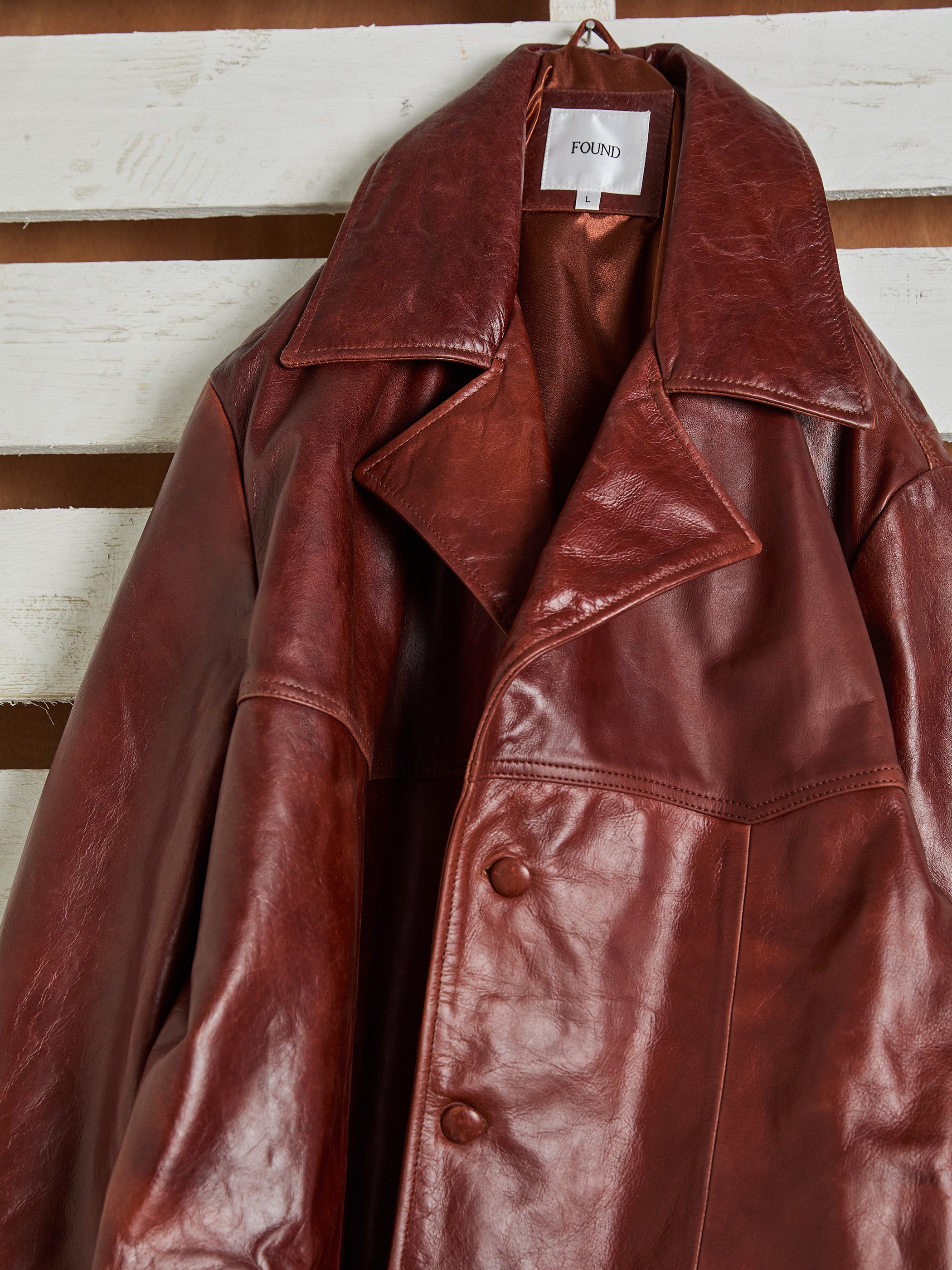 The FOUND Claret Western Oversized Leather Blazer, unisex with large lapels and a double-breasted front, elegantly hangs against a white wooden backdrop, merging classic style with contemporary charm.