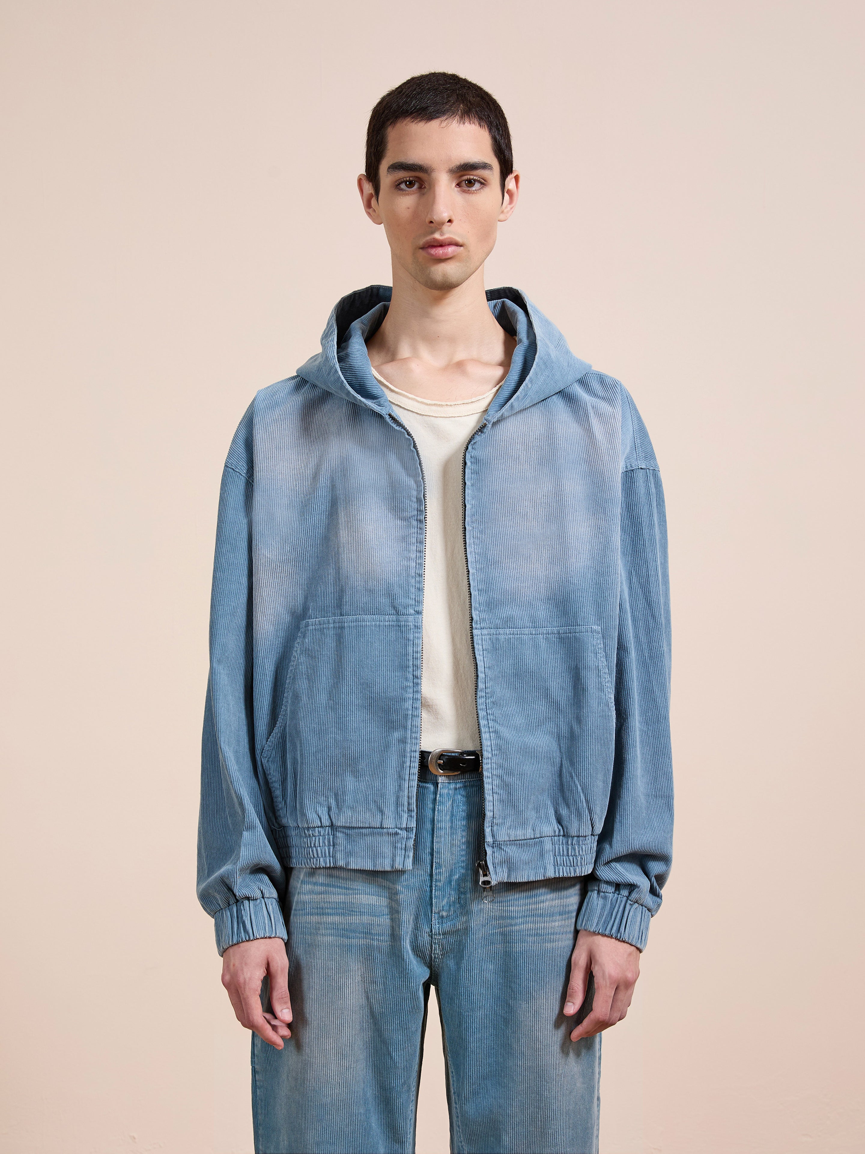 A person wears the FOUND Corduroy Zip Up Hoodie in light blue over a white shirt with matching blue jeans, standing against a plain beige background.