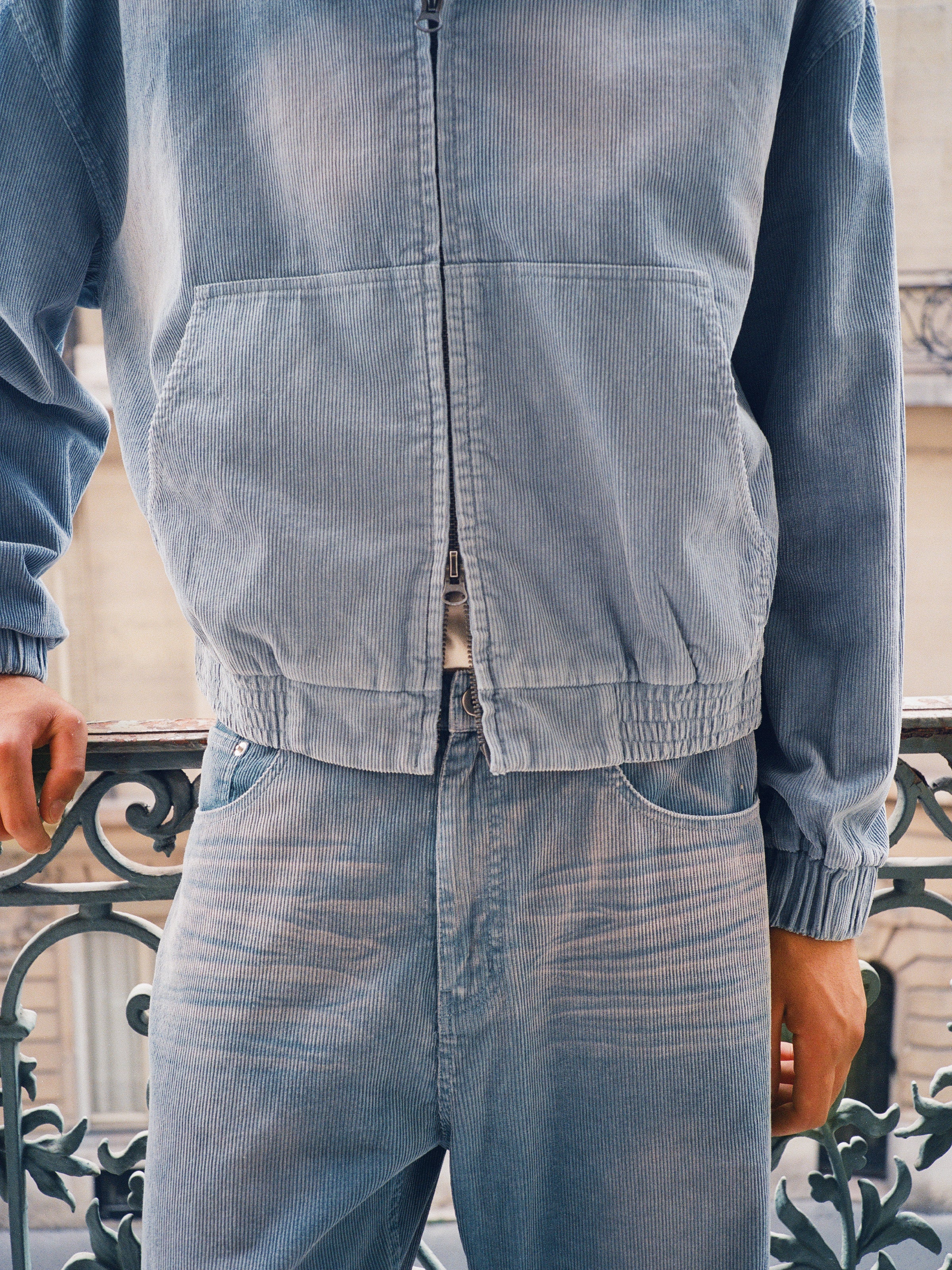 A person stands by a decorative metal railing wearing the FOUND Corduroy Zip Up Hoodie in light blue, paired with matching pants.
