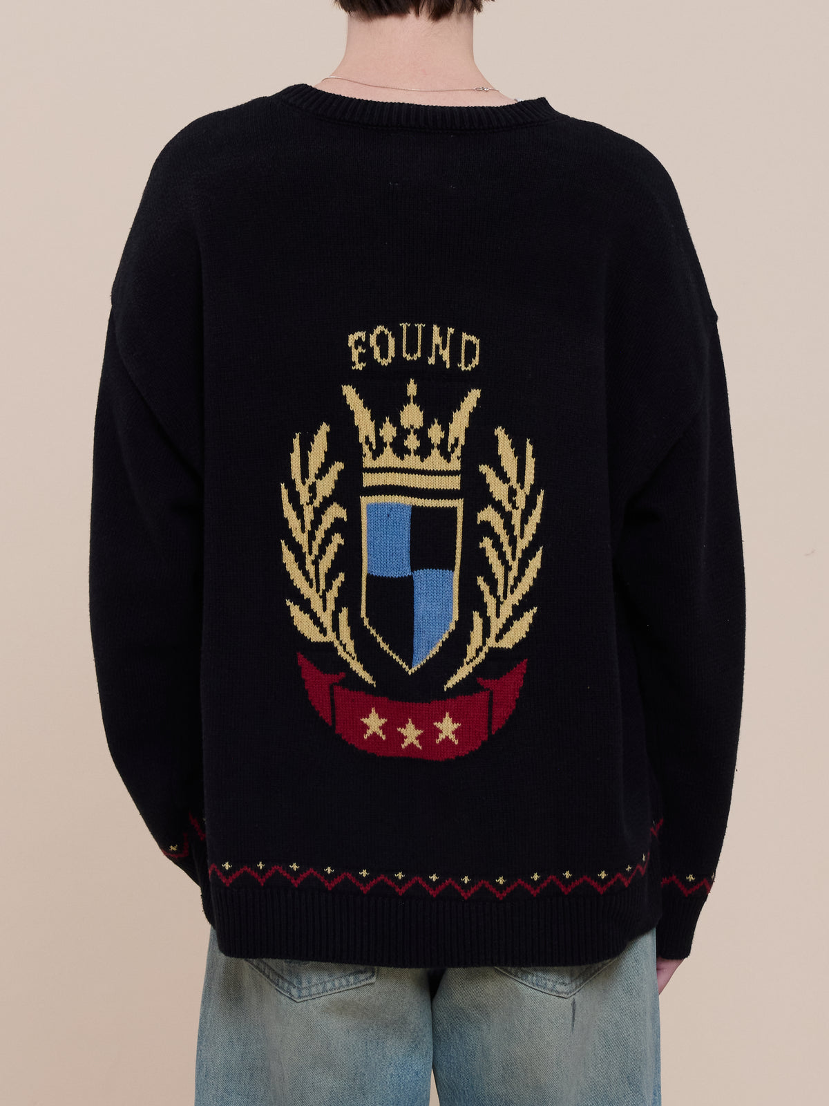 Person in a black Crest Sweater by FOUND, featuring a gold laurel, crown, shield, and red stars on the back—an eye-catching collegiate knit with blue jeans.