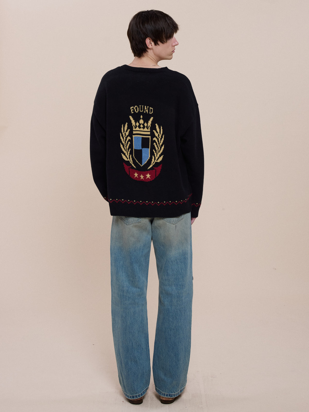 Person faces away in light blue wide-leg jeans and a black FOUND Crest Sweater with laurel branches, red stars, and “FOUND,” set against a beige background.