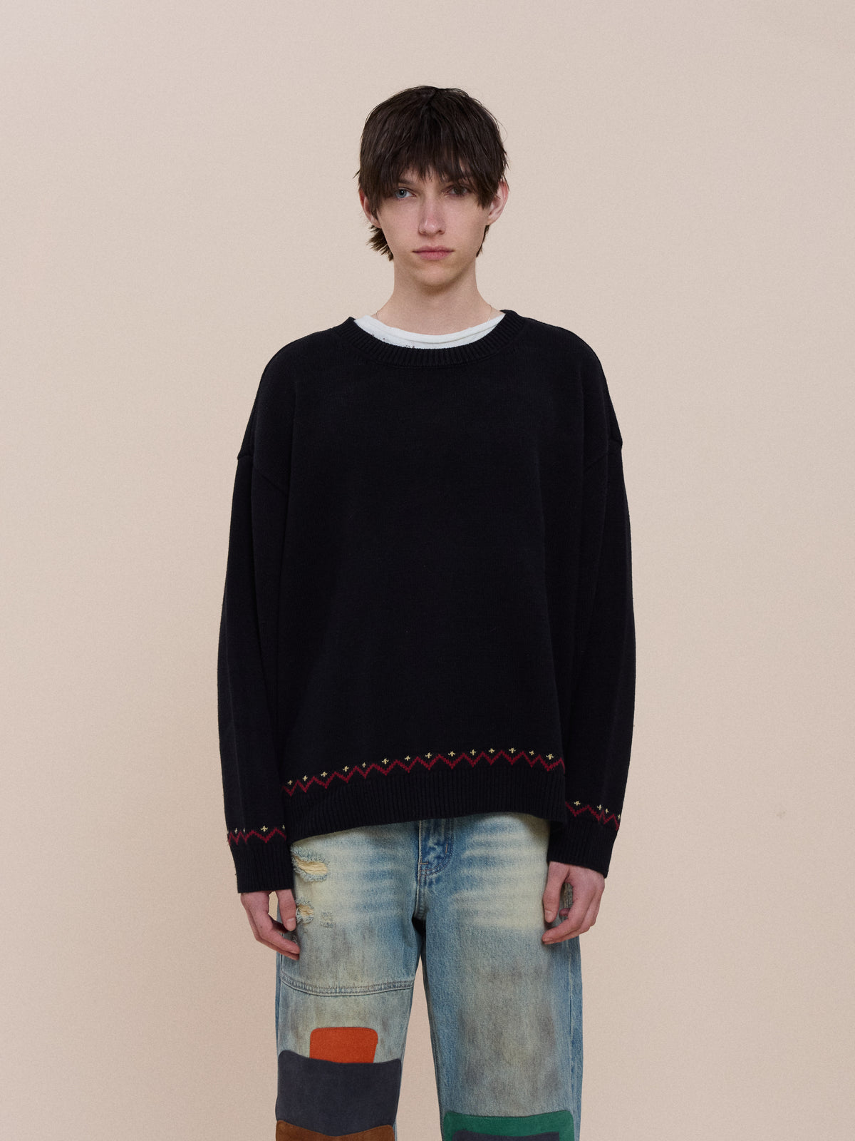 Person with short brown hair wears the FOUND Crest Sweater and light blue patchwork jeans, standing against a plain beige background facing the camera.
