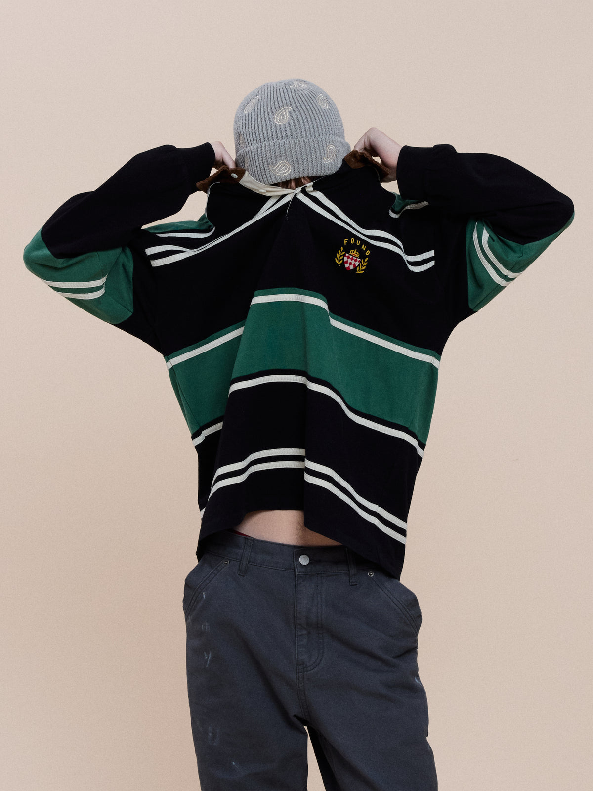 Model wearing the FOUND Crestfield Rugby in green and black stripes with gray pants, lifts the shirt over their head, facing away from the camera.