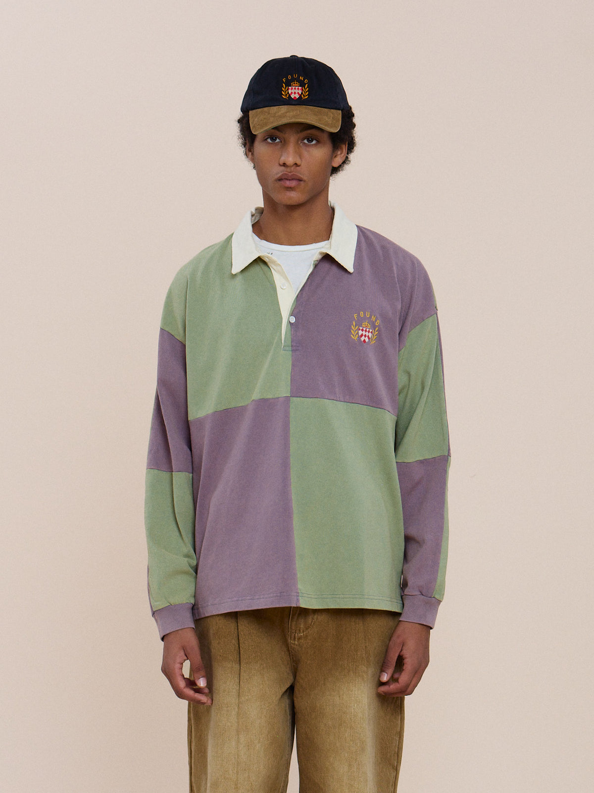 A person in a green and purple Crestfield Rugby by FOUND, tan pants, and a black cap stands against a plain light background, looking at the camera.