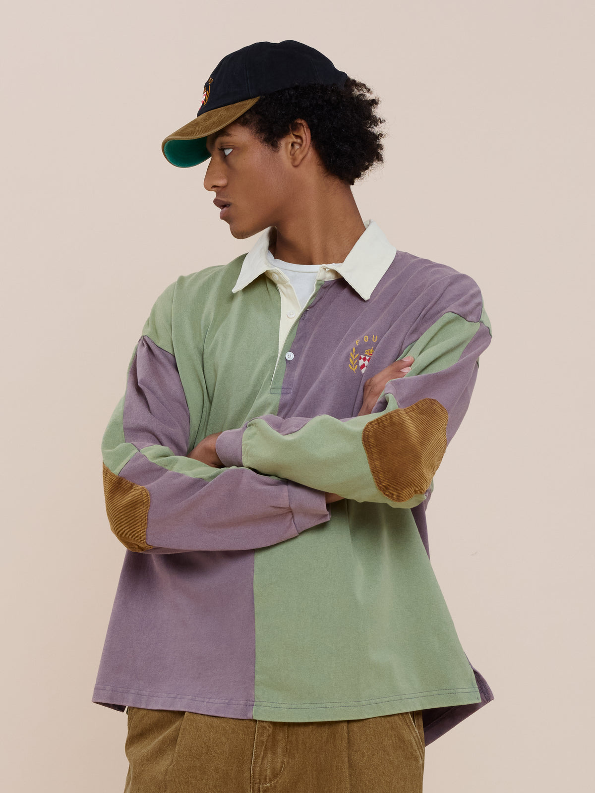 A person wears the FOUND Crestfield Rugby—a vintage-inspired, two-tone green and purple shirt with brown elbow patches—paired with a black cap and brown pants.