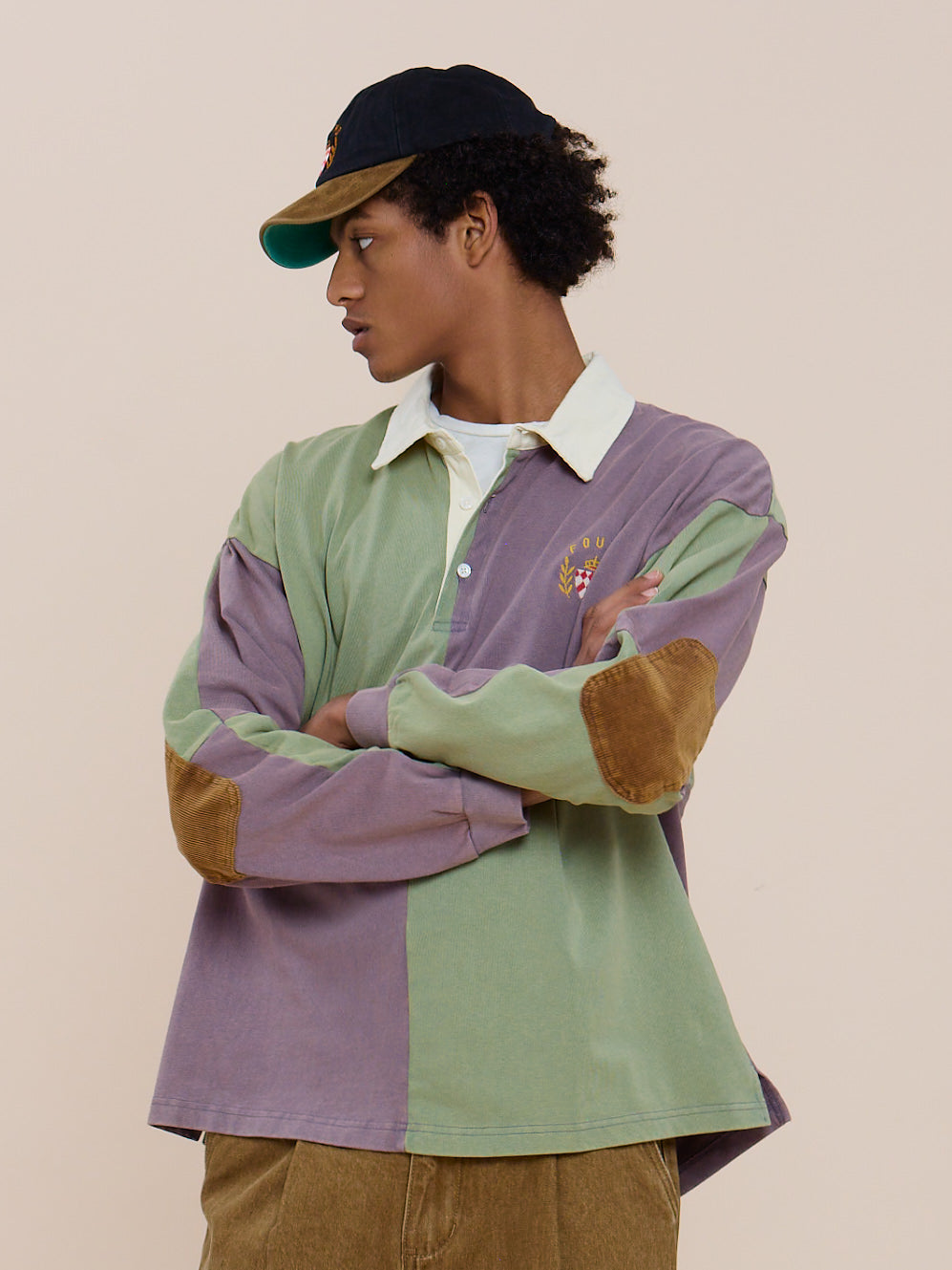 Person wearing the FOUND Crestfield Rugby—a green and purple color-block rugby shirt with brown elbow patches—tan pants, and a black cap, arms crossed, looking to the side.
