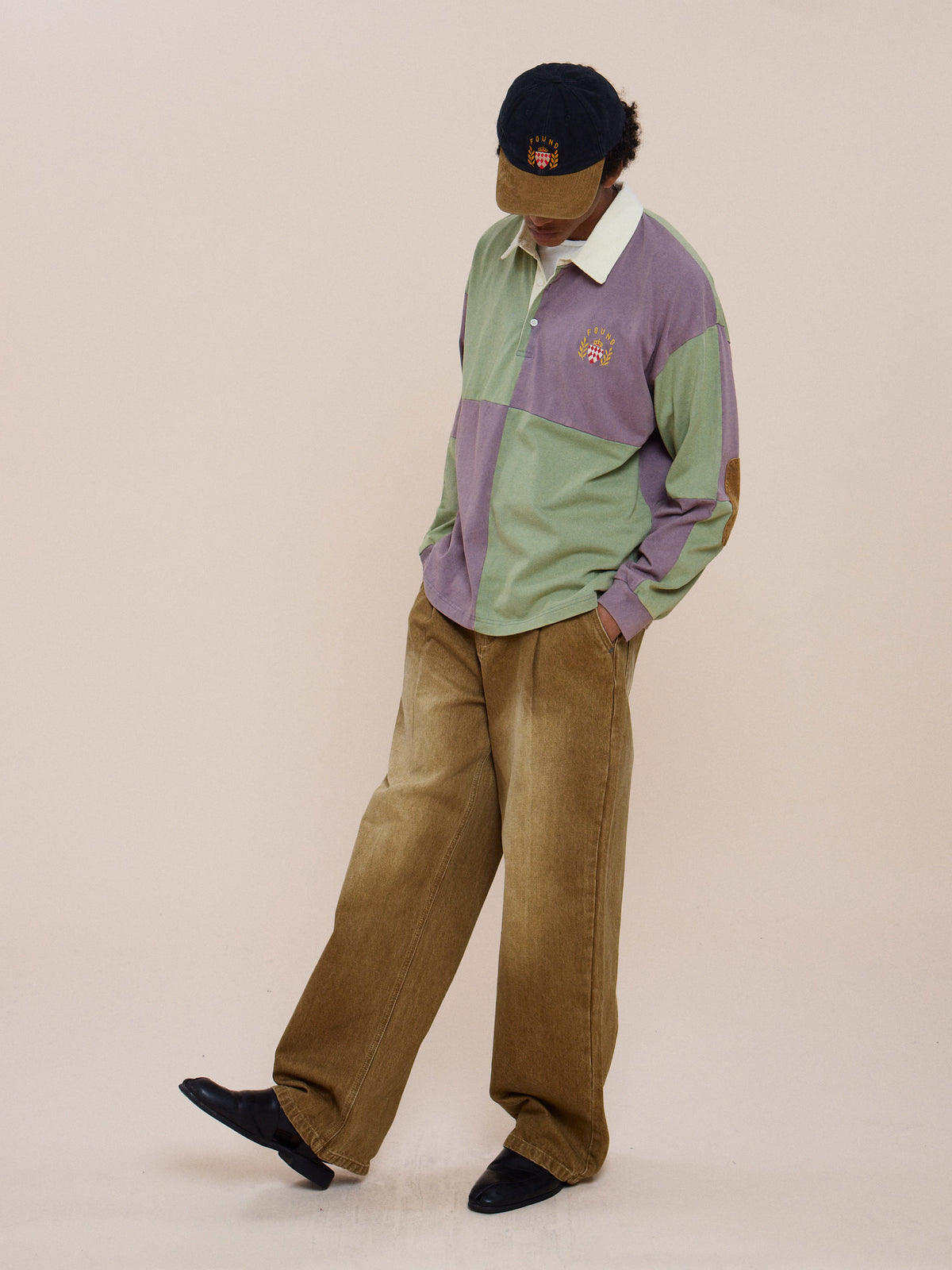 Person in the FOUND Crestfield Rugby, wide tan pants, black shoes, and a dark cap stands looking down against a plain background.