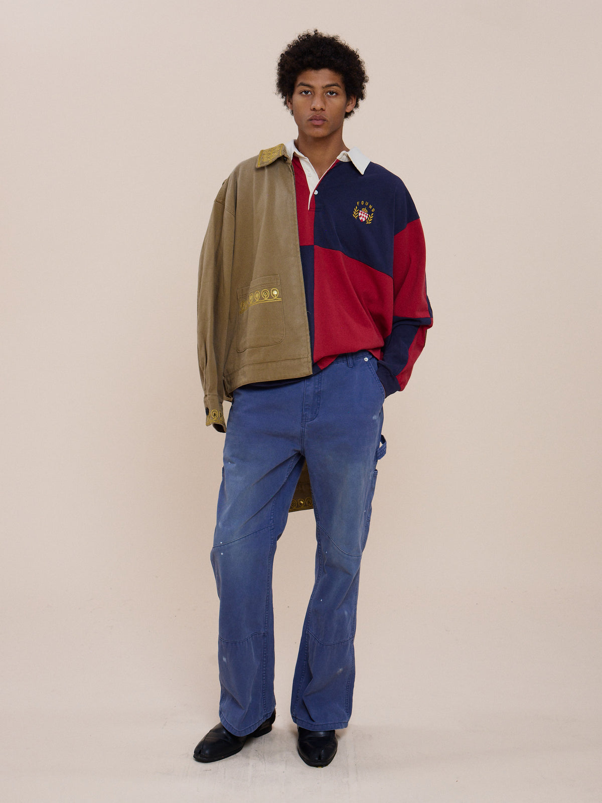 A person stands against a plain backdrop wearing a tan jacket draped over one shoulder, the Crestfield Rugby by FOUND, blue pants, and black shoes.