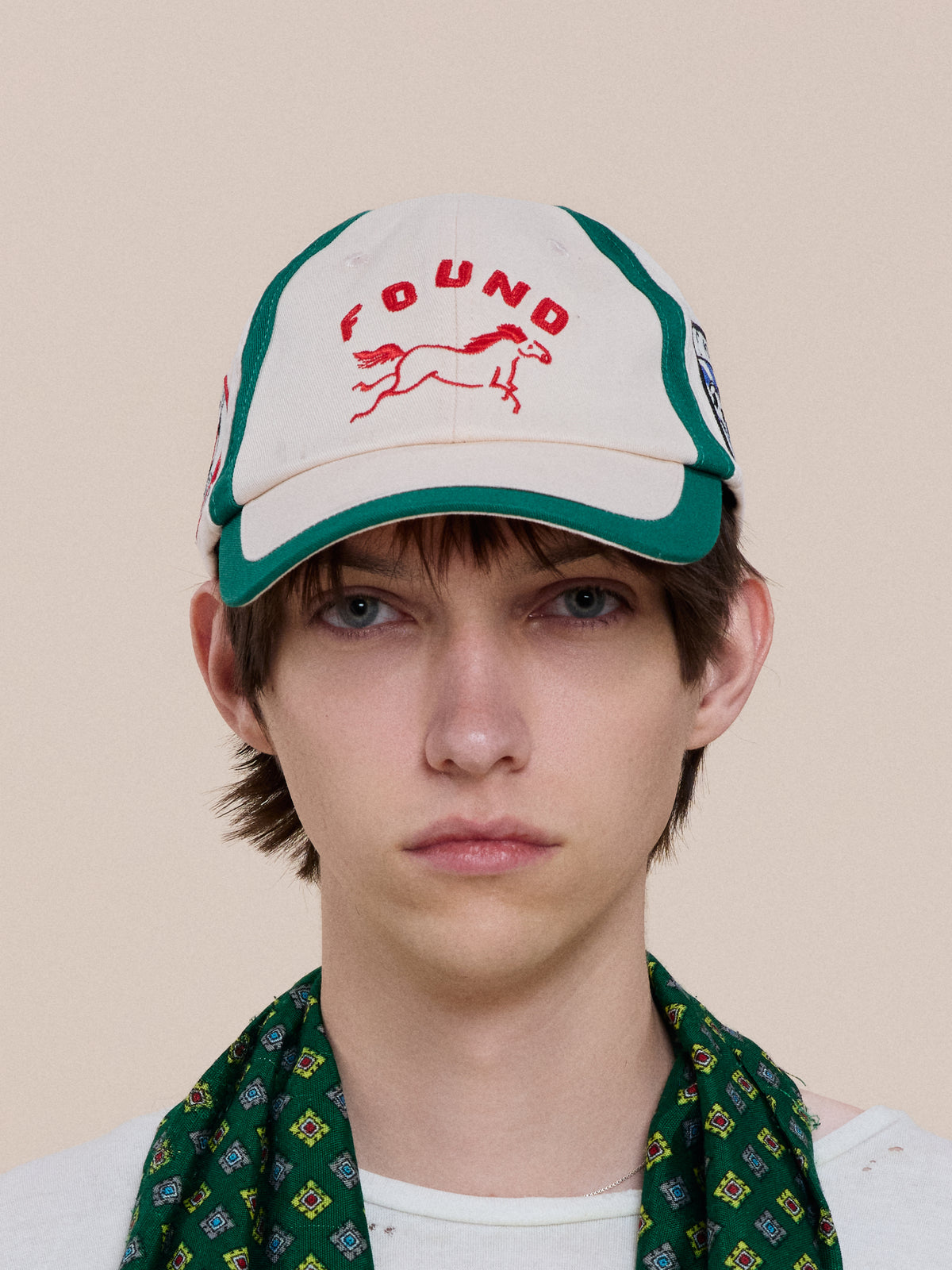 A person with short brown hair wears the Crestline Racing Cap by Found (white/green, horse graphic) and a green patterned scarf, facing forward against a plain background.