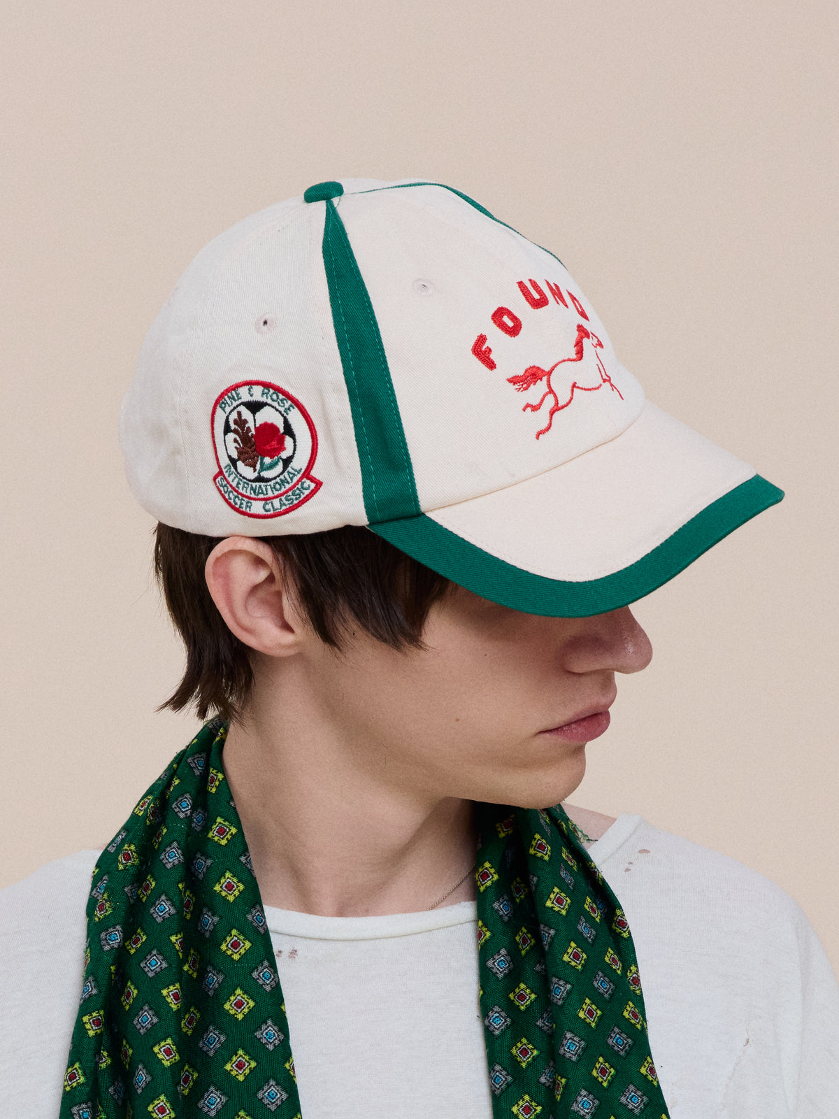 Person wearing a white and green Crestline Racing Cap by Found with red text and patch, looking to the side, paired with a green patterned scarf.