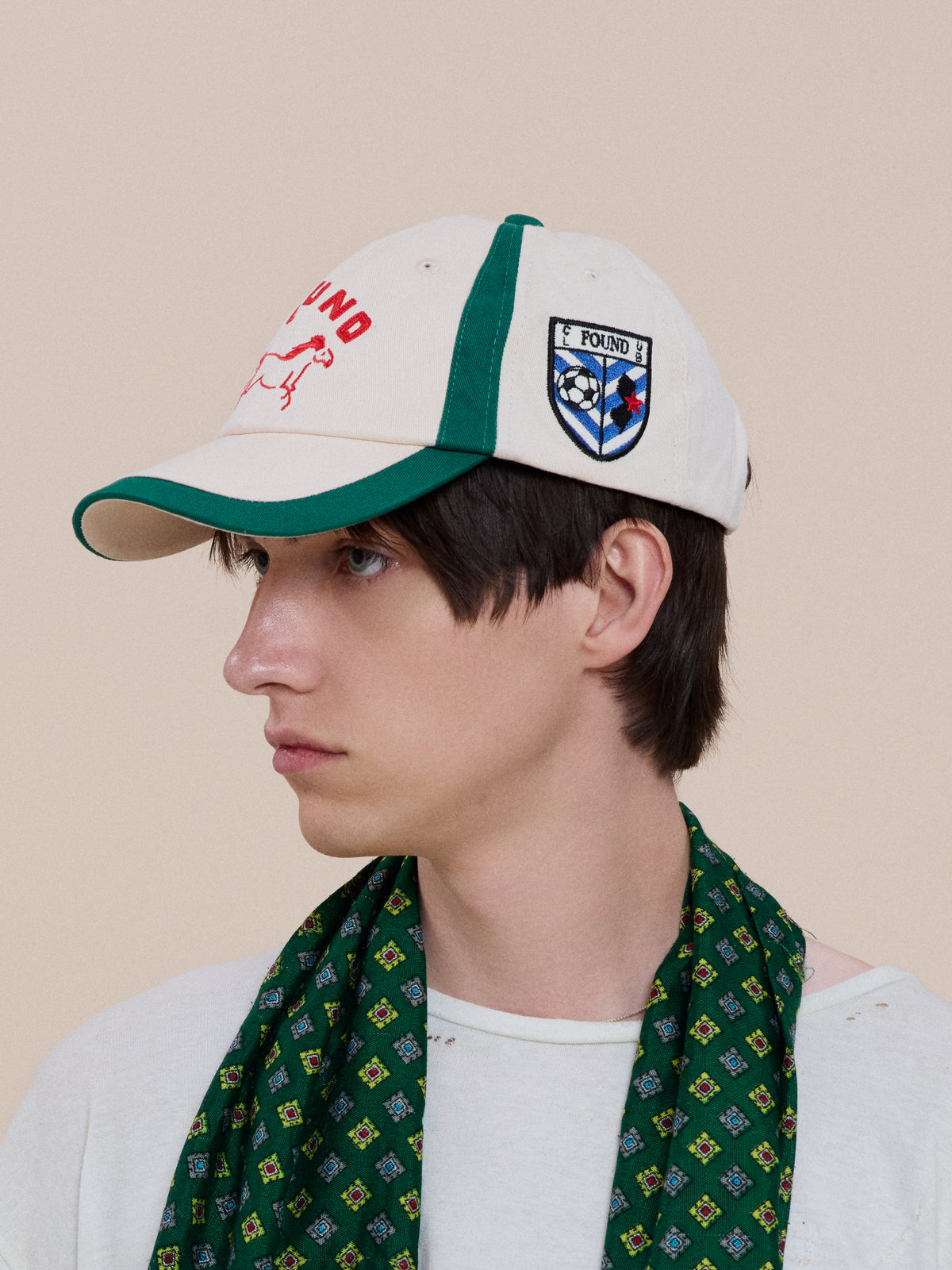 Person in a Crestline Racing Cap by Found (white and green, “FOUND” text, crest), white shirt, and green patterned scarf, facing left.