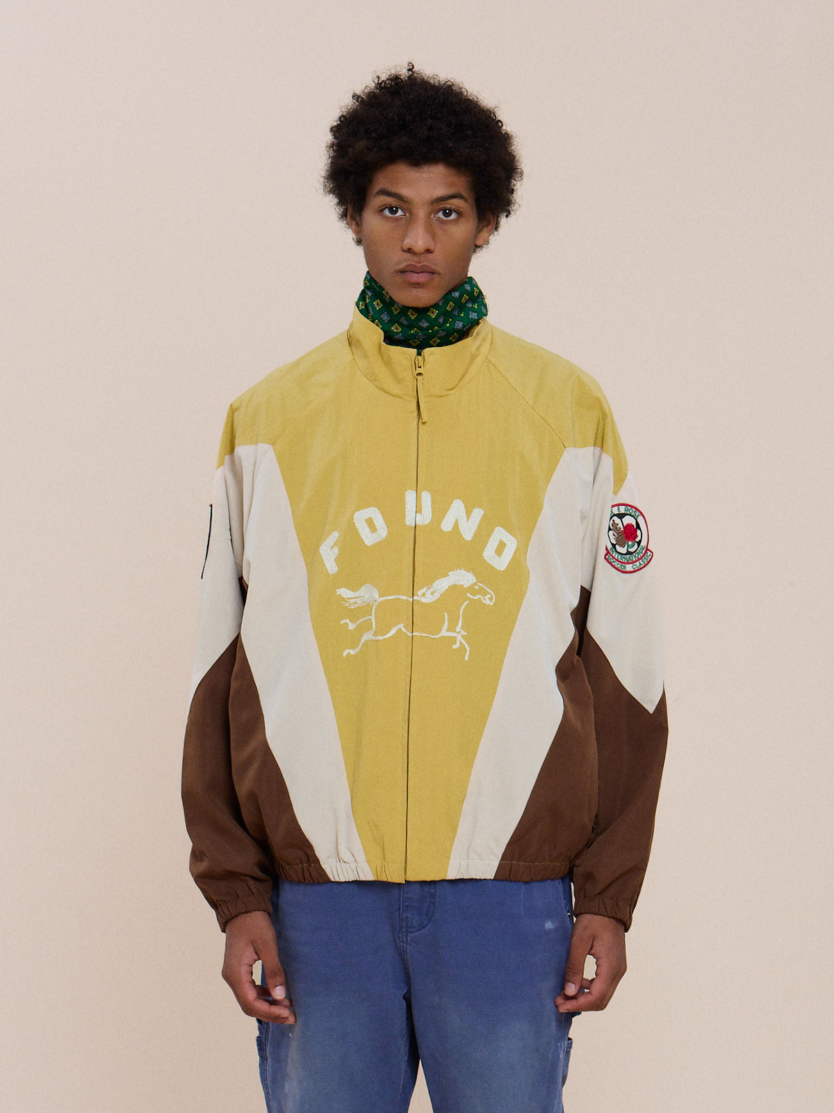 A person wears the Crestline Sport Zip Jacket by FOUND—yellow, brown, and white with a horse graphic—blue jeans, and a green scarf against a plain background.