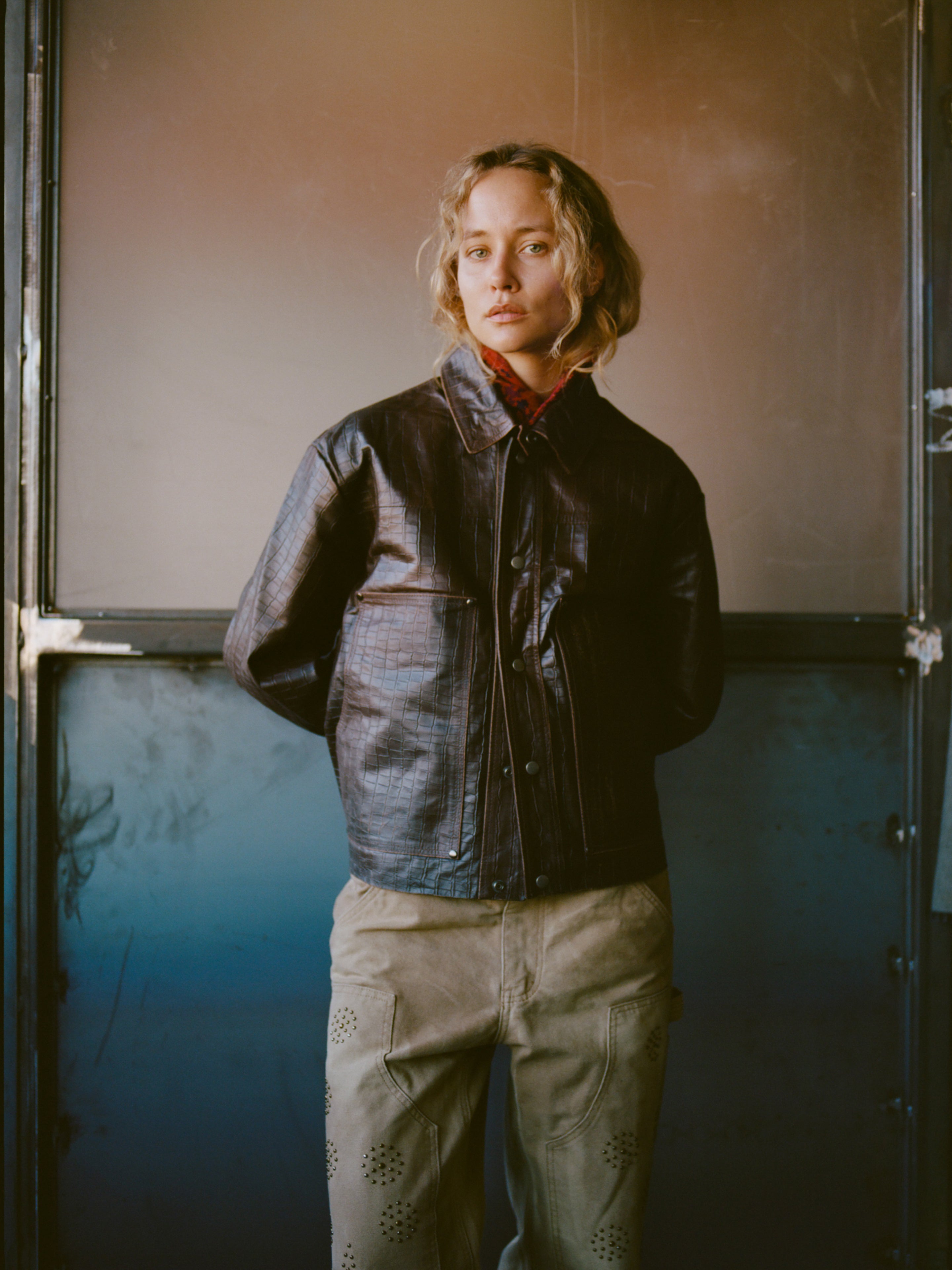 A person with shoulder-length curly hair wears the FOUND Distressed Croc Embossed Leather Work Jacket and beige pants, standing indoors against a metallic, gradient-toned background.