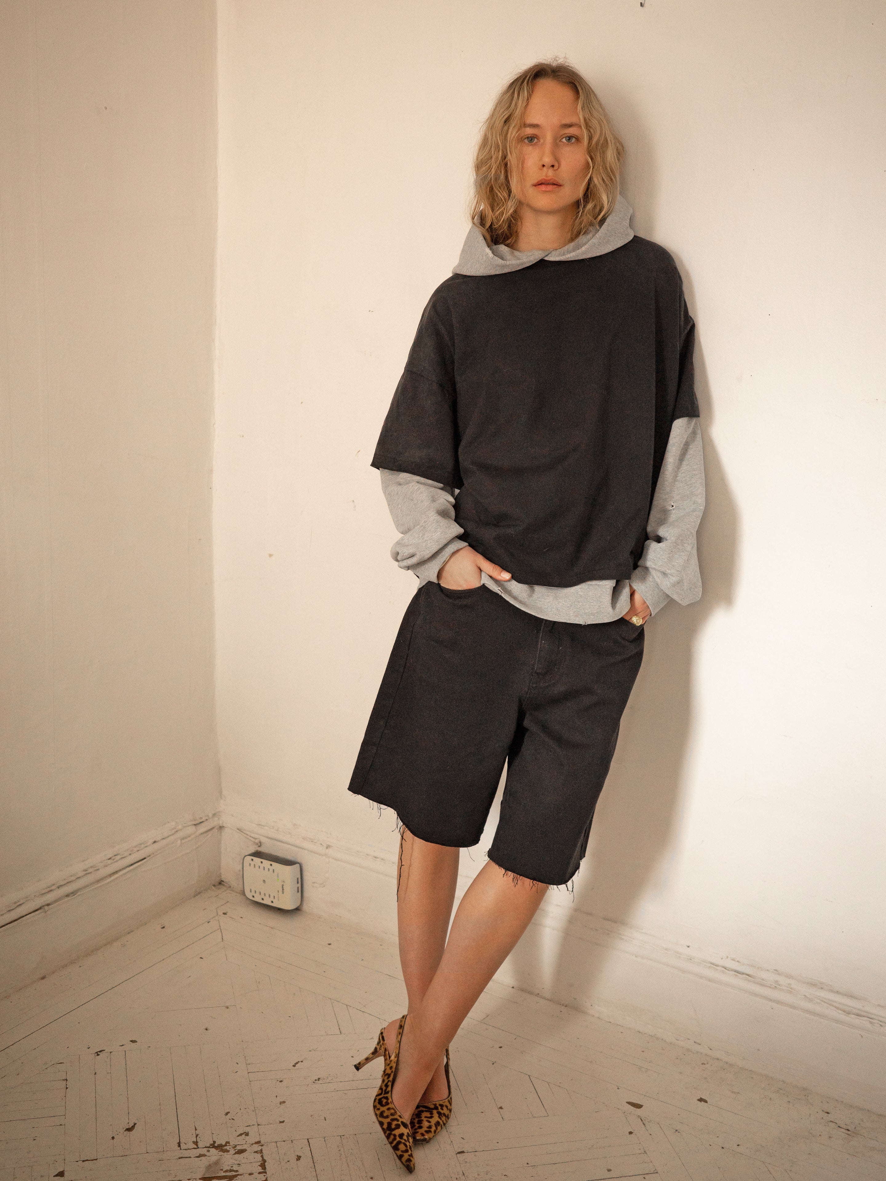 A person stands in a corner against a plain wall, sporting a vintage look with the Found Double Layer Hoodie, shorts, and leopard print heels.