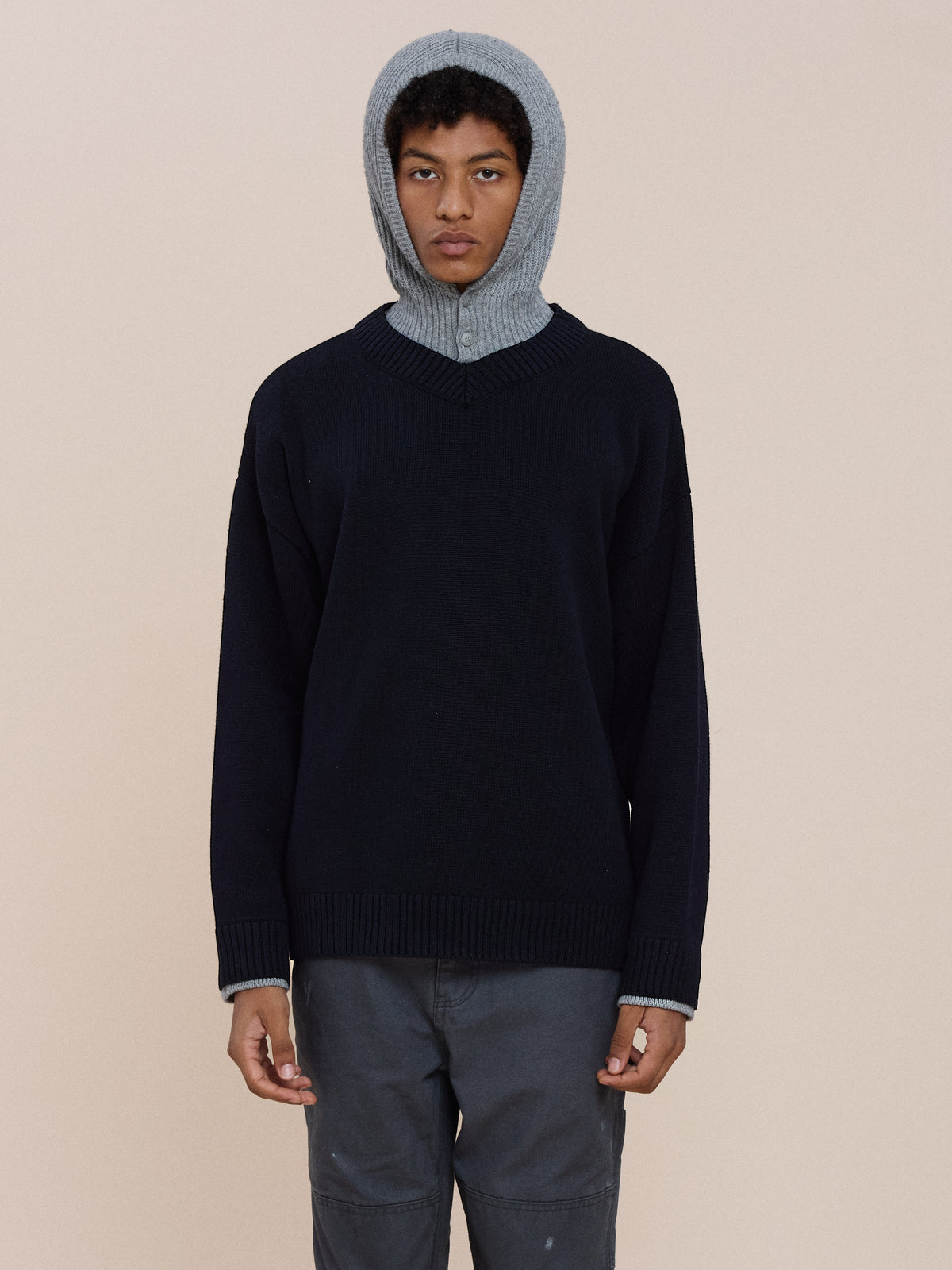 A person wears the FOUND Double Layer V Neck Knit Sweater over a gray hoodie with gray pants, standing against a plain beige background.