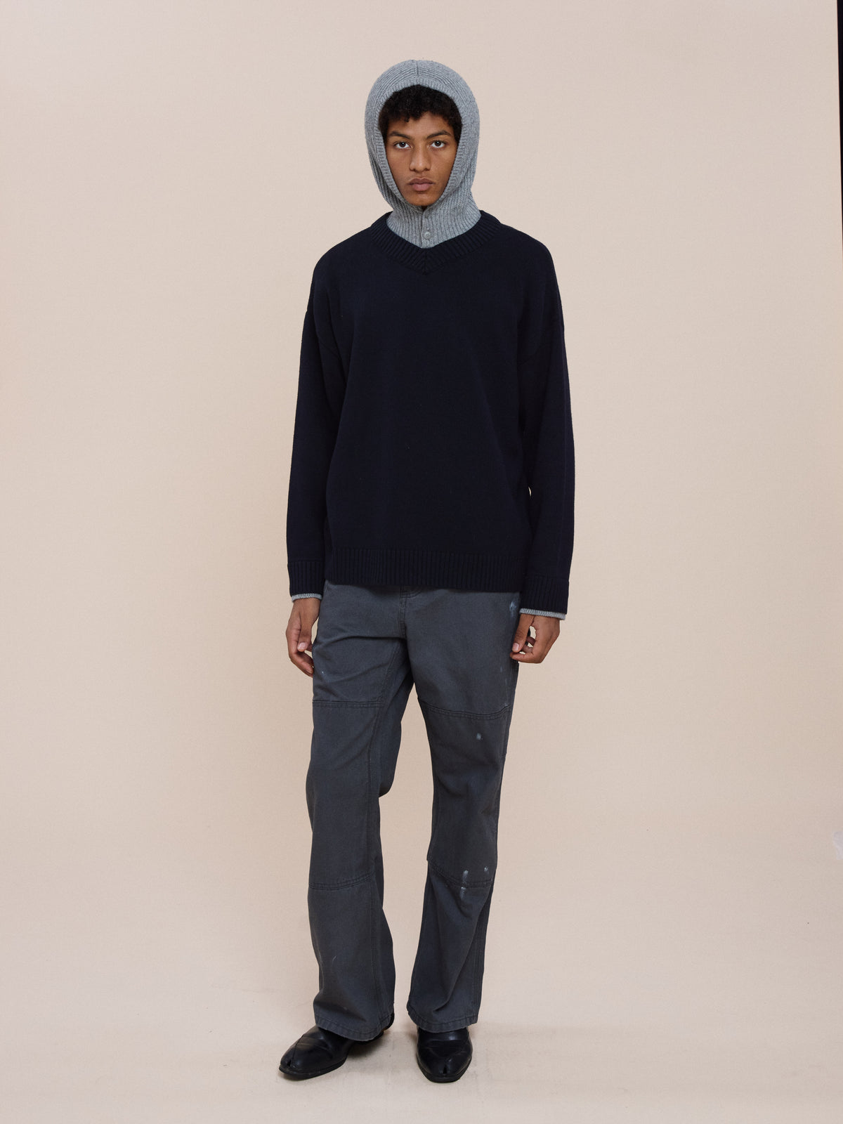 Person wears a gray Double Layer V Neck Knit Sweater by FOUND, gray pants, and black shoes, standing against a plain beige background.