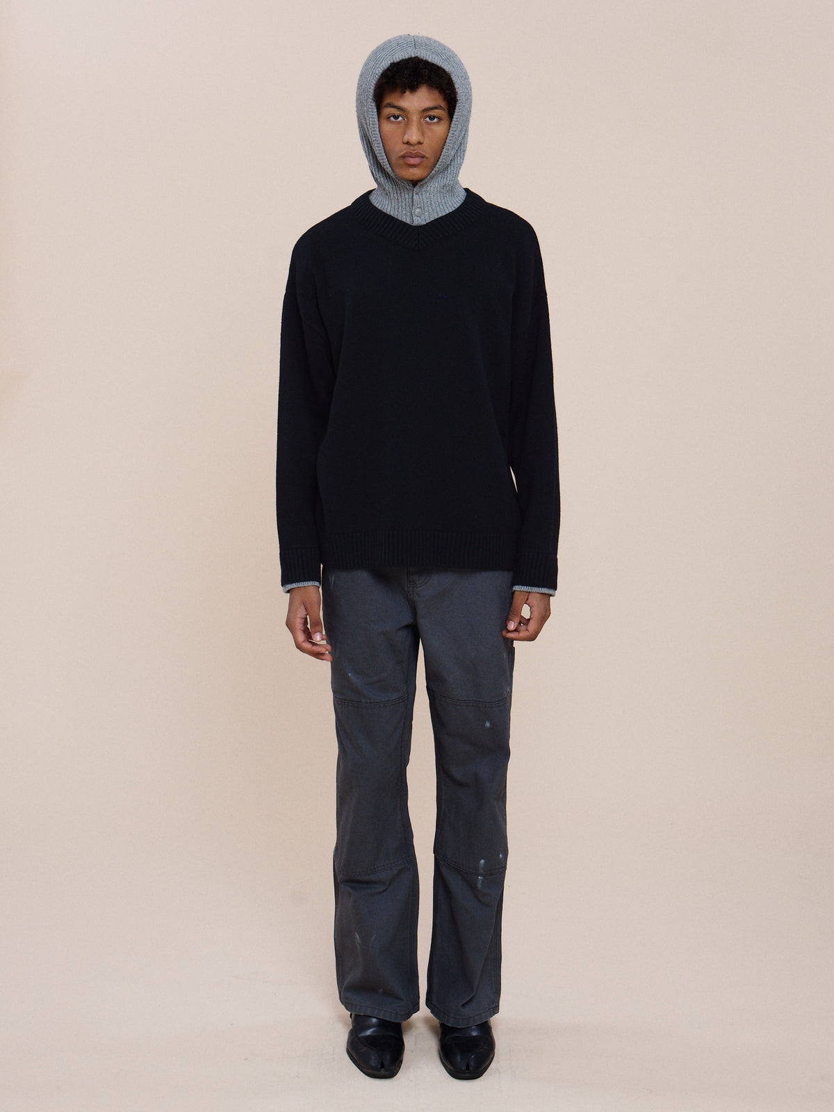Person standing against a plain background, wearing FOUND Double Layer V Neck Knit Sweater, gray pants, black shoes, and a gray knit hood.