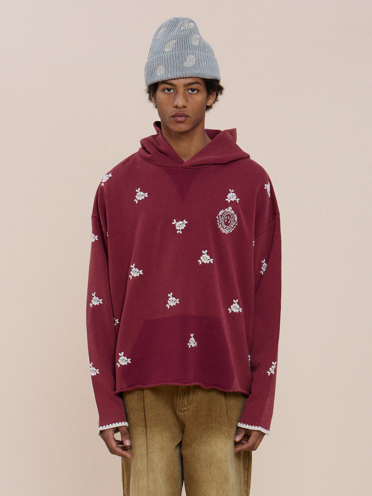 Someone wearing tan pants, a light blue knit hat, and the FOUND Embroidered Motif Hoodie stands against a plain beige background.