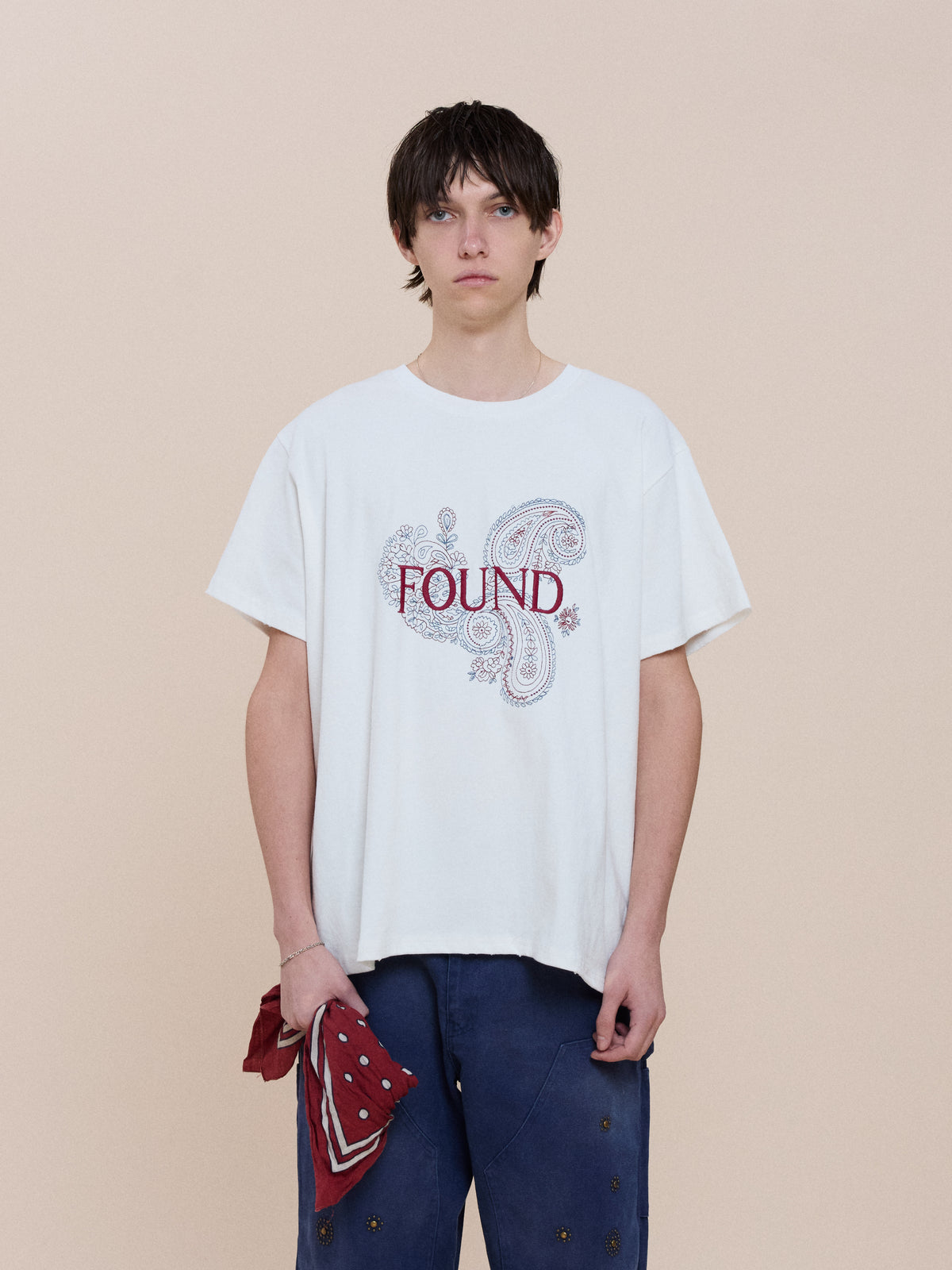 Person with straight dark hair wears the FOUND Embroidered Paisley Tee in white and blue pants, holding a red bandana print against a plain beige background.